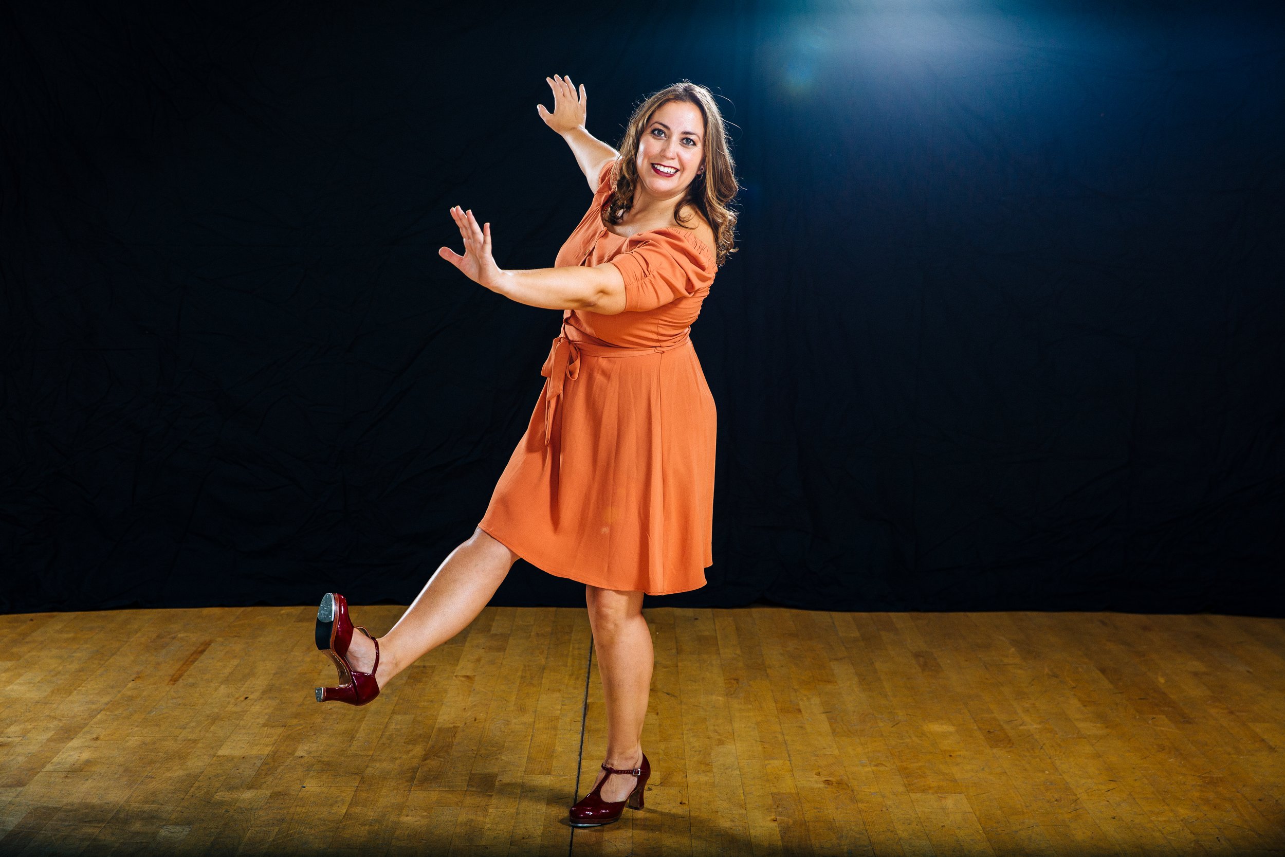 A woman in an orange dress and red heels posing playfully on a wooden floor against a black background.