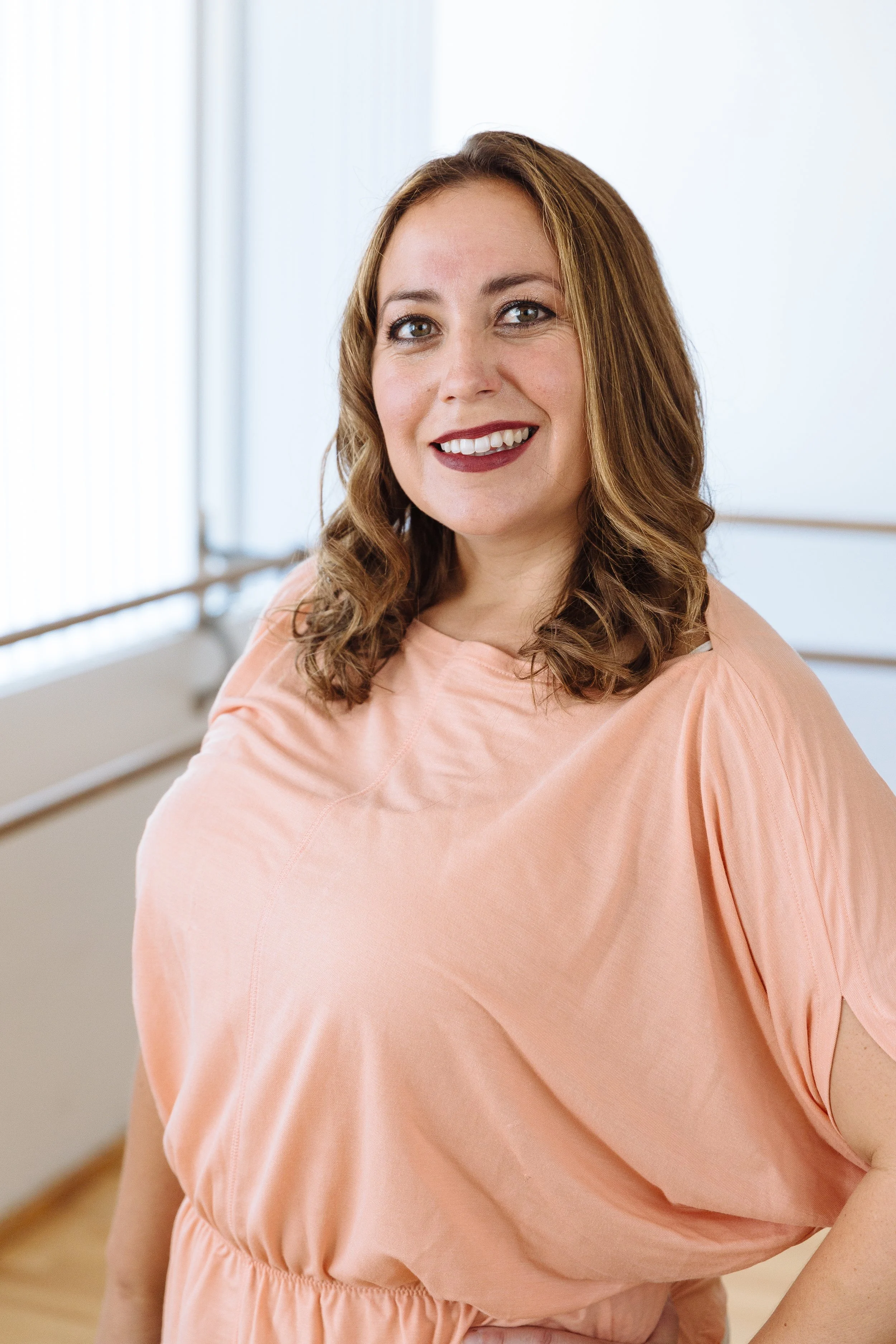A smiling woman with medium-length wavy brown hair wearing a peach-colored top inside a bright room.