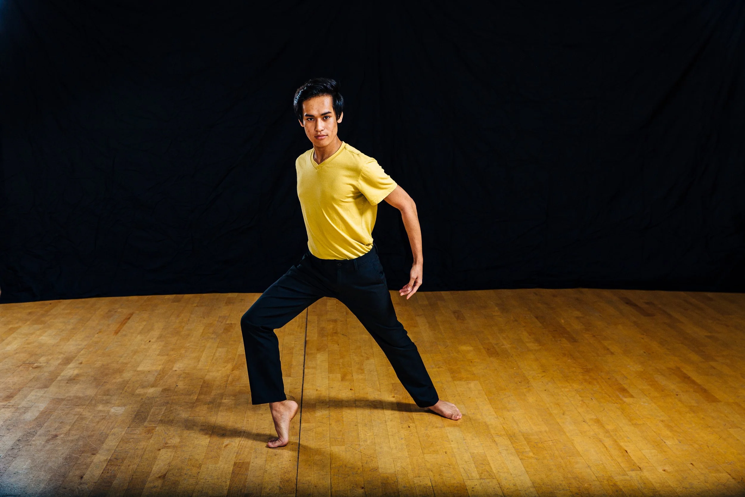 A young man in a yellow t-shirt and black pants demonstrates martial arts stance in a studio with a wooden floor and black backdrop.
