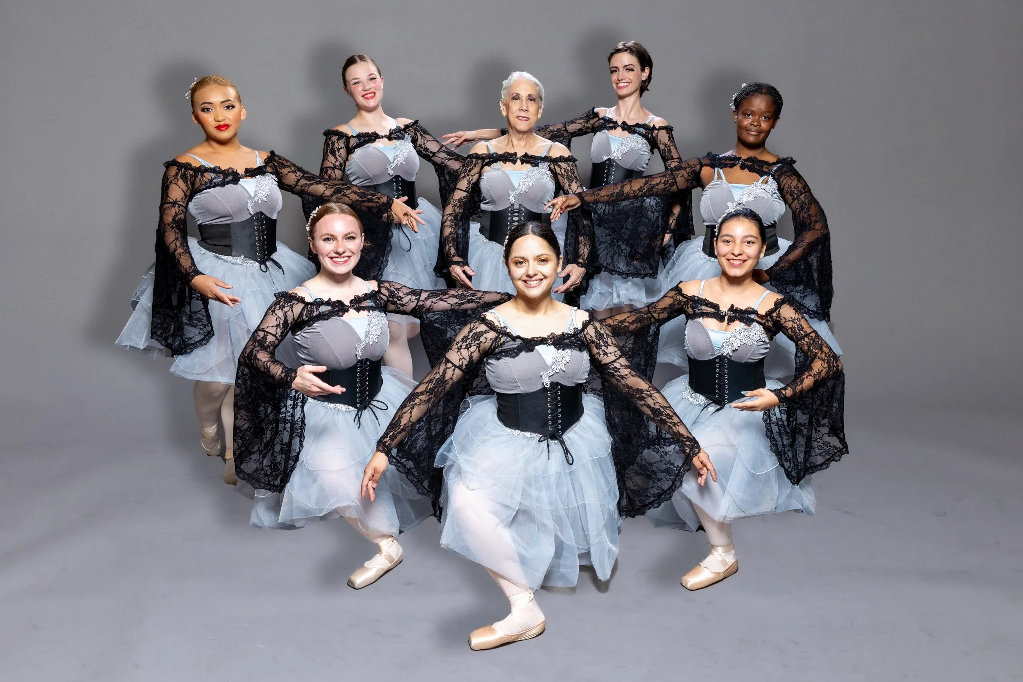 Group of ten ballet dancers in costumes posing in a studio with a gray background.