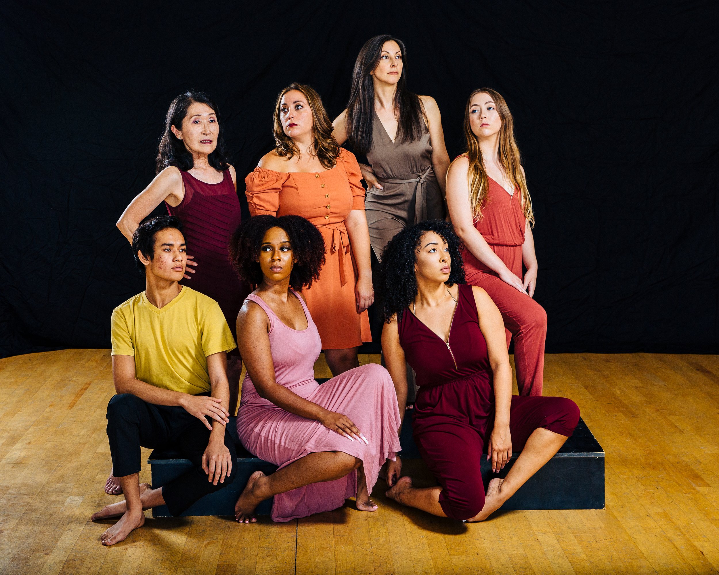 Group of eight women of diverse ages and ethnicities, posing together in a studio with a black backdrop and wooden floor, some sitting, others standing.