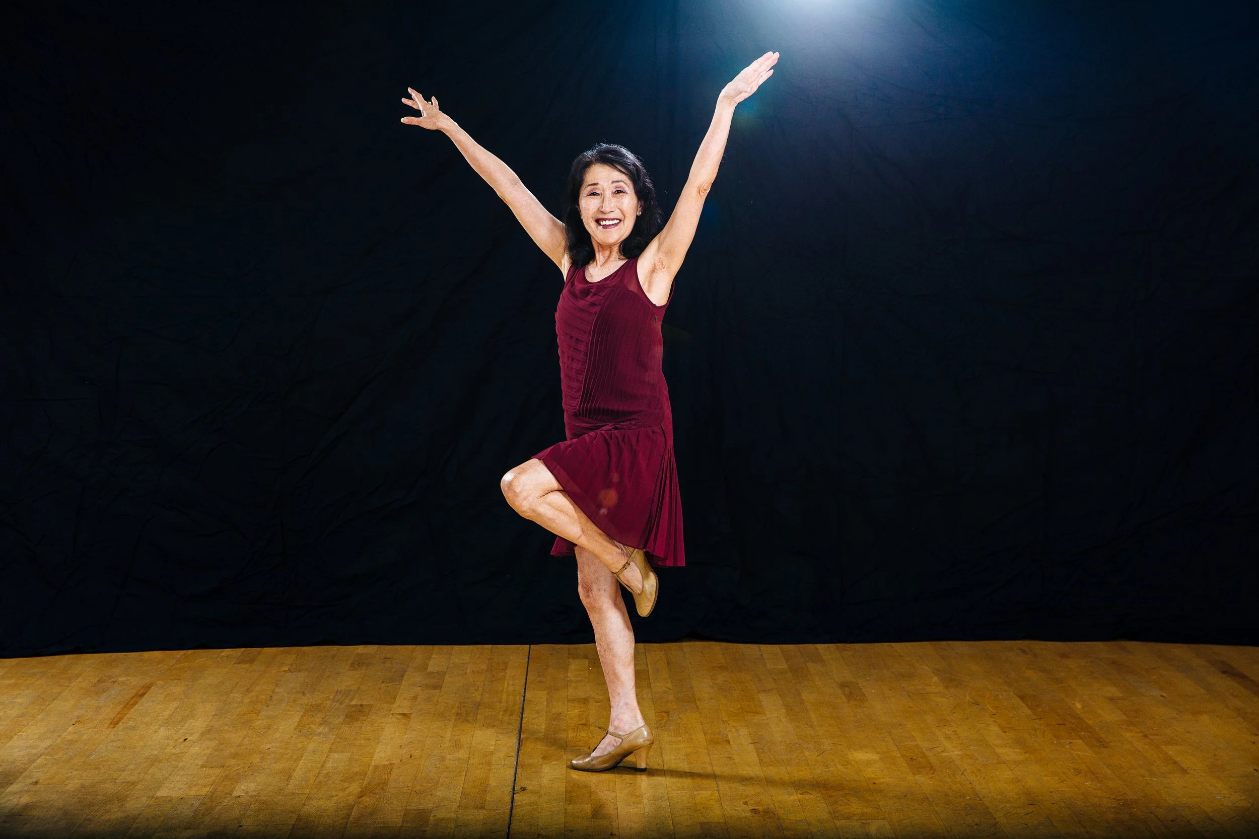 A woman in a red dress and high heels dancing on a wooden floor with arms raised and one leg bent, smiling in front of a dark background with a spotlight overhead.