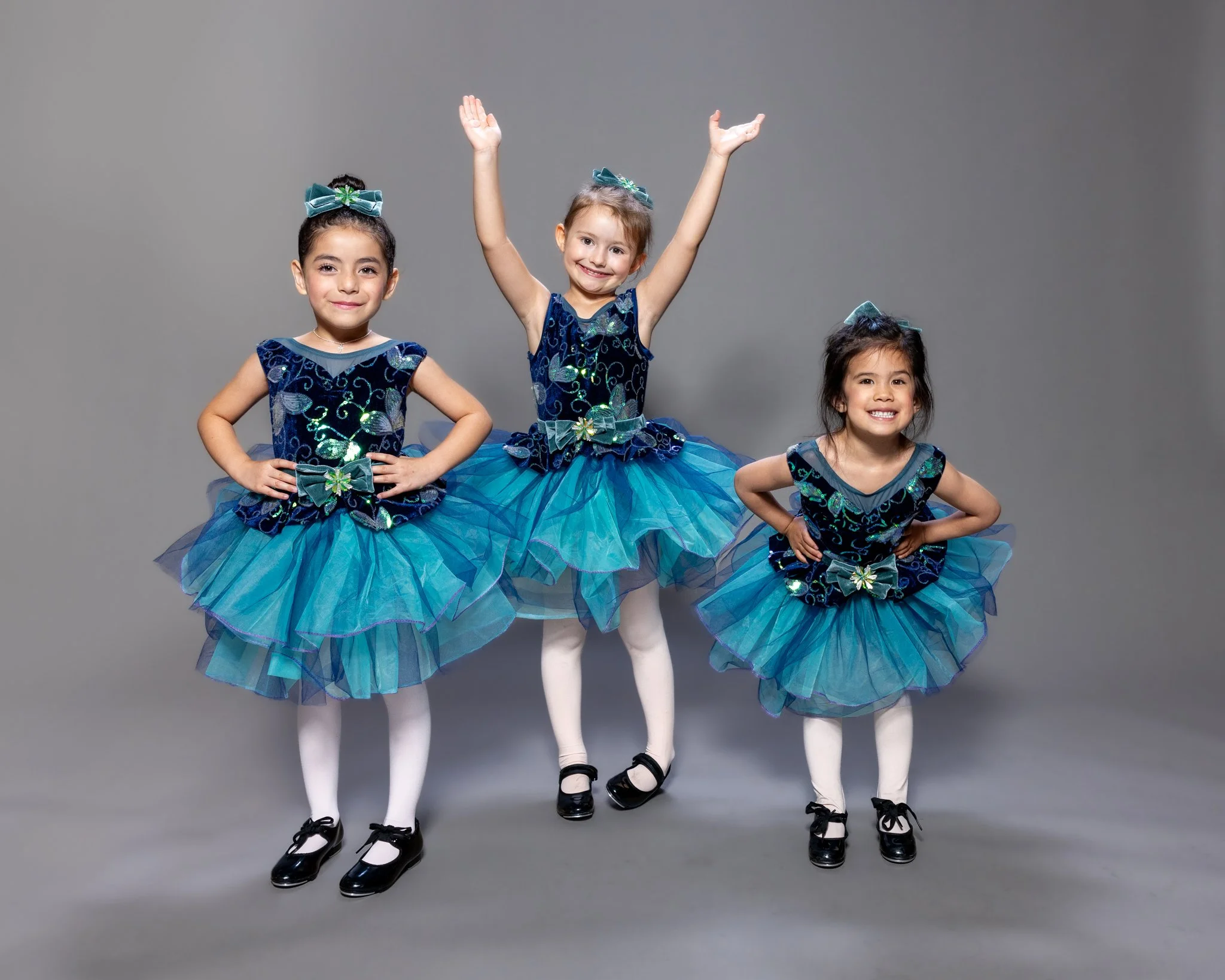 Three young girls in matching blue dance costumes, posing with hands on hips or raised, smiling against a gray background.