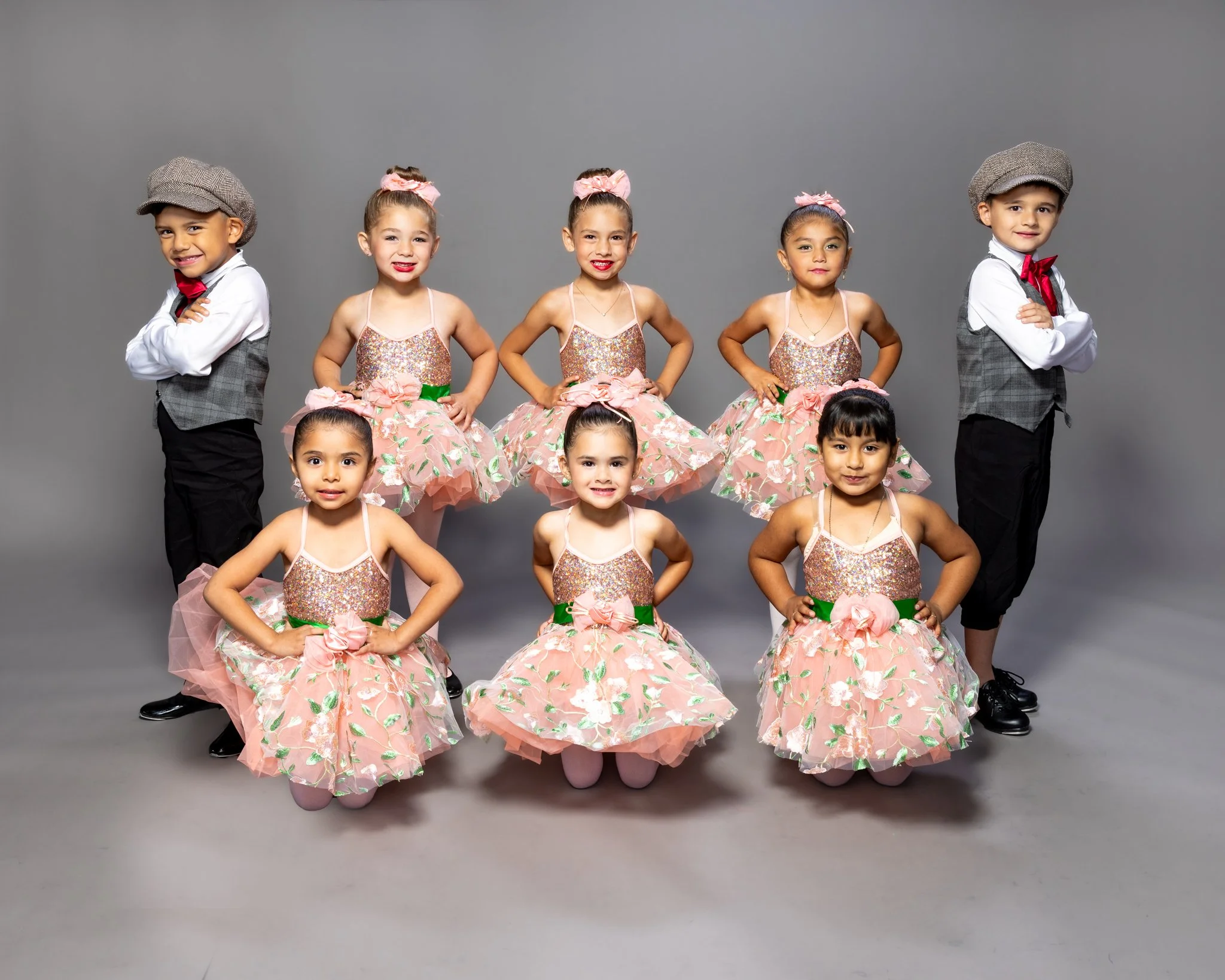 Group of seven young children, four girls in pink floral dance costumes and three boys in vintage dress, standing against a gray background.