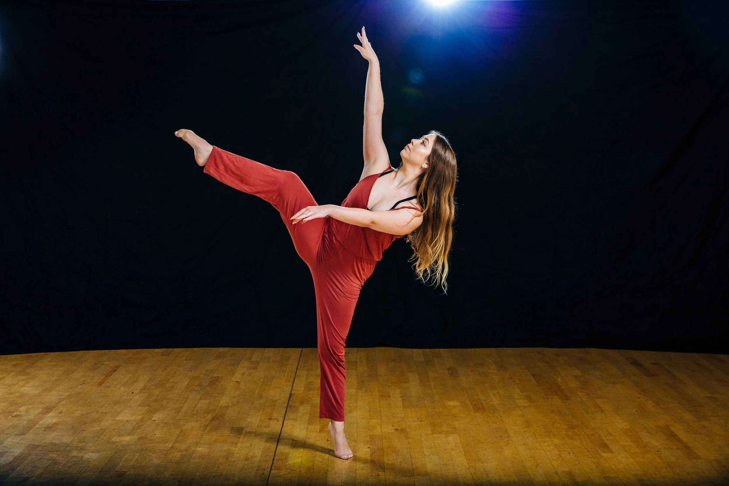 A woman dancing or performing a pose in a studio with a black background and wooden floor, wearing a red outfit.