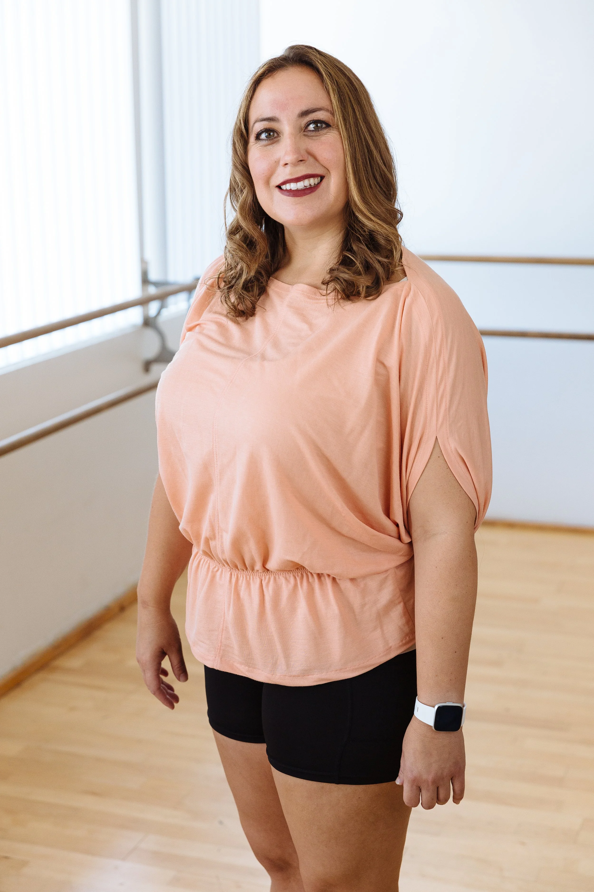 A woman with long brown curly hair wearing a peach-colored top, black shorts, and a white smartwatch standing in a dance studio with wooden floors and ballet barre rails.