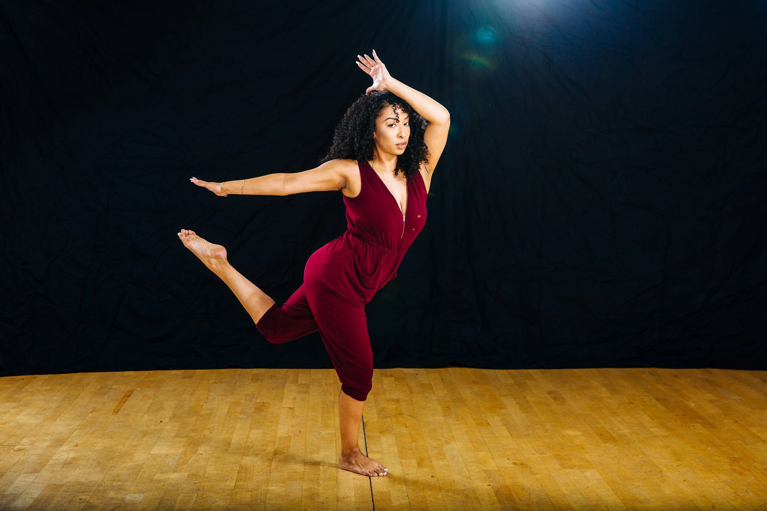 A woman with curly hair wearing a burgundy jumpsuit performing a yoga pose on a wooden floor against a black backdrop.