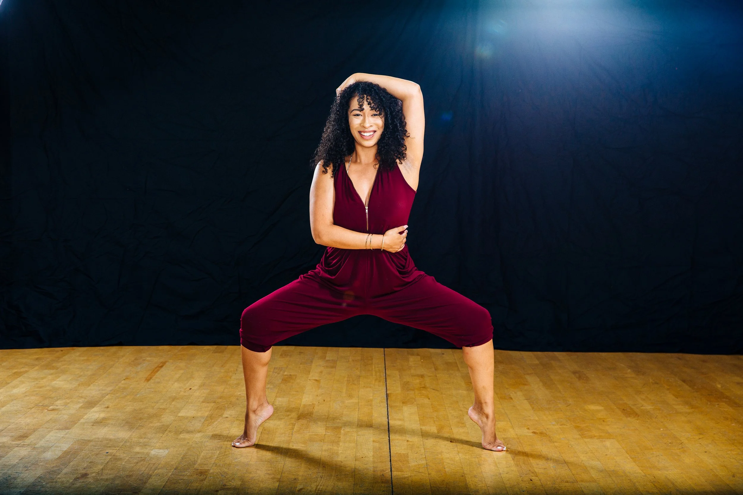 Woman in a red jumpsuit doing a yoga pose with legs wide apart and one arm up, smiling, on a wooden floor against a dark background.