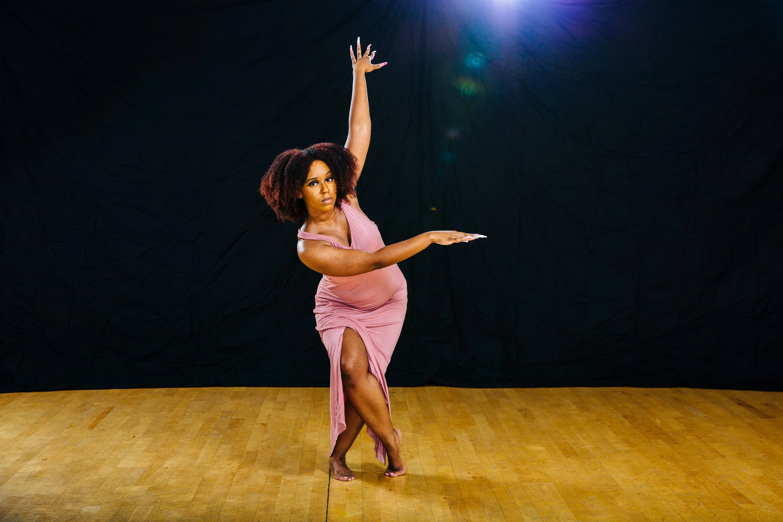 A woman with curly hair wearing a pink dress and performing a dance move on a wooden floor with a black background.