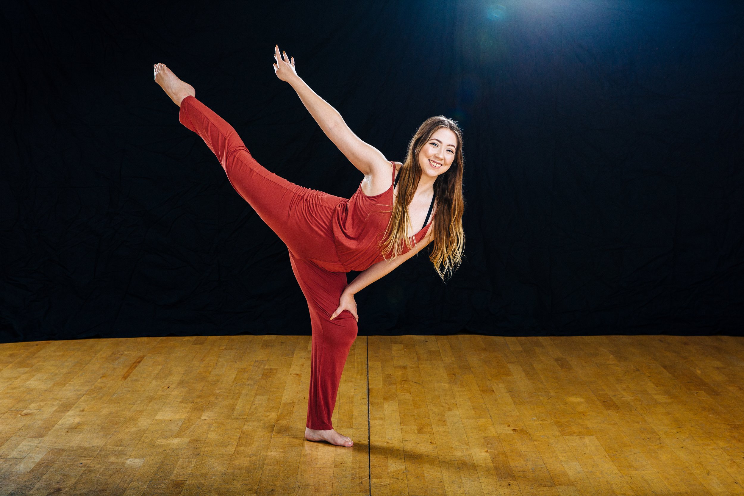 Young woman with long hair in a red sleeveless outfit performs a yoga pose on a wooden floor against a dark background, smiling.