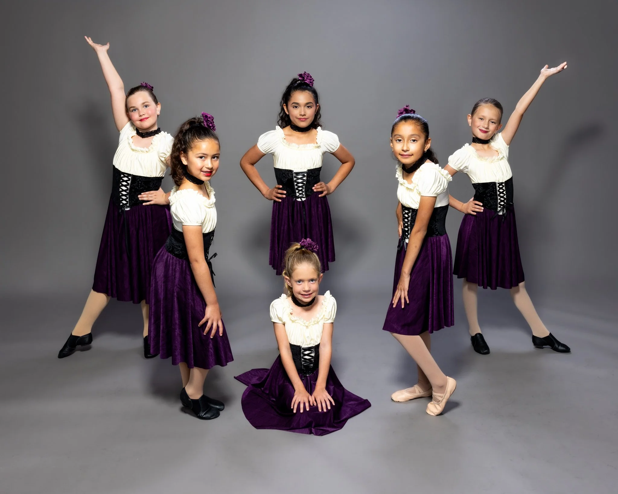 Six young girls in matching dance costumes pose against a gray backdrop. The costumes include purple skirts, white blouses, and black corset-style belts; some have purple accessories in their hair. The girls display various dance poses, with one knee