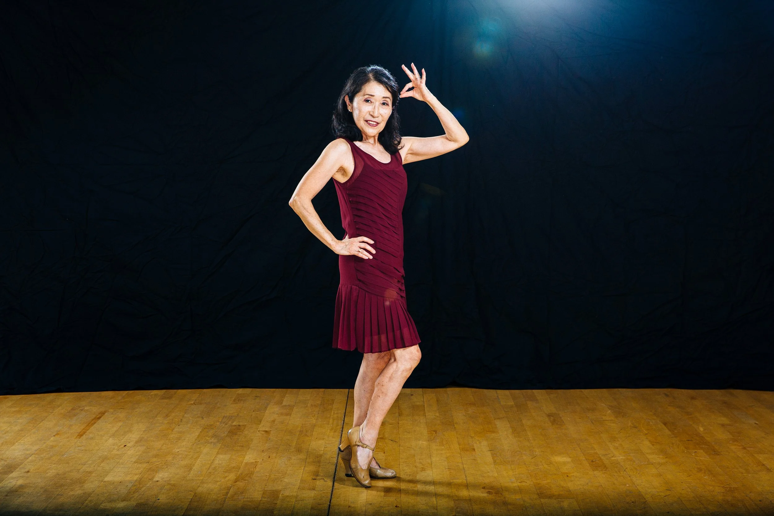 An older woman in a red dress and high heels dancing on a wooden floor against a black background.