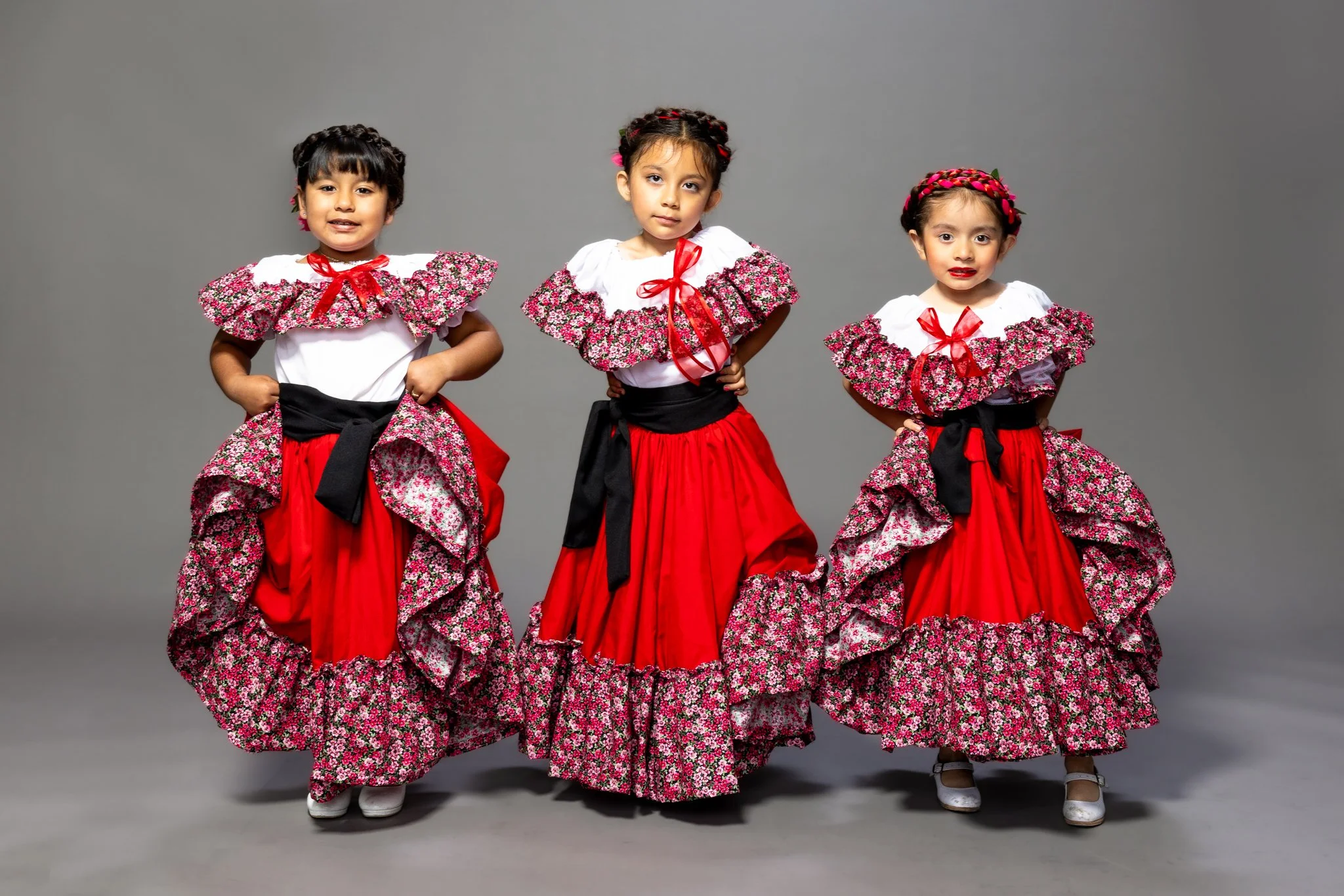 Three young girls dressed in traditional Mexican folkloric dresses with red skirts, floral ruffled tops, and red ribbons in their hair, standing against a gray background.