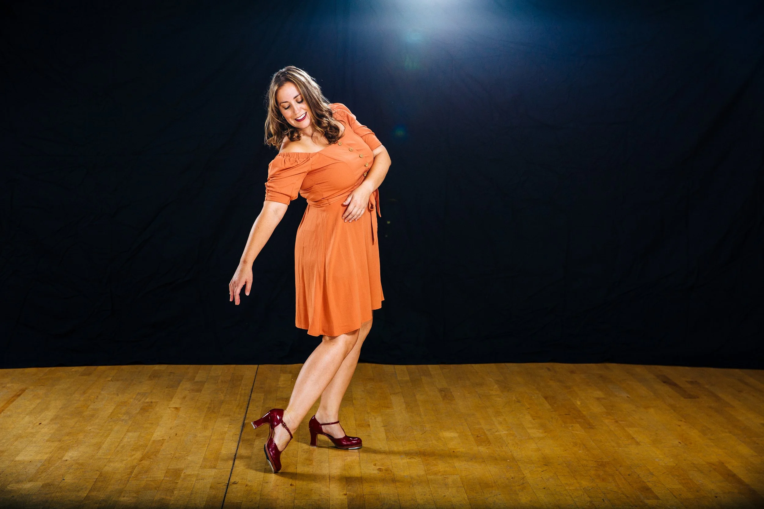 A woman with brown hair wearing an orange dress and red high heels dancing on a wooden floor against a dark background with a light source overhead.