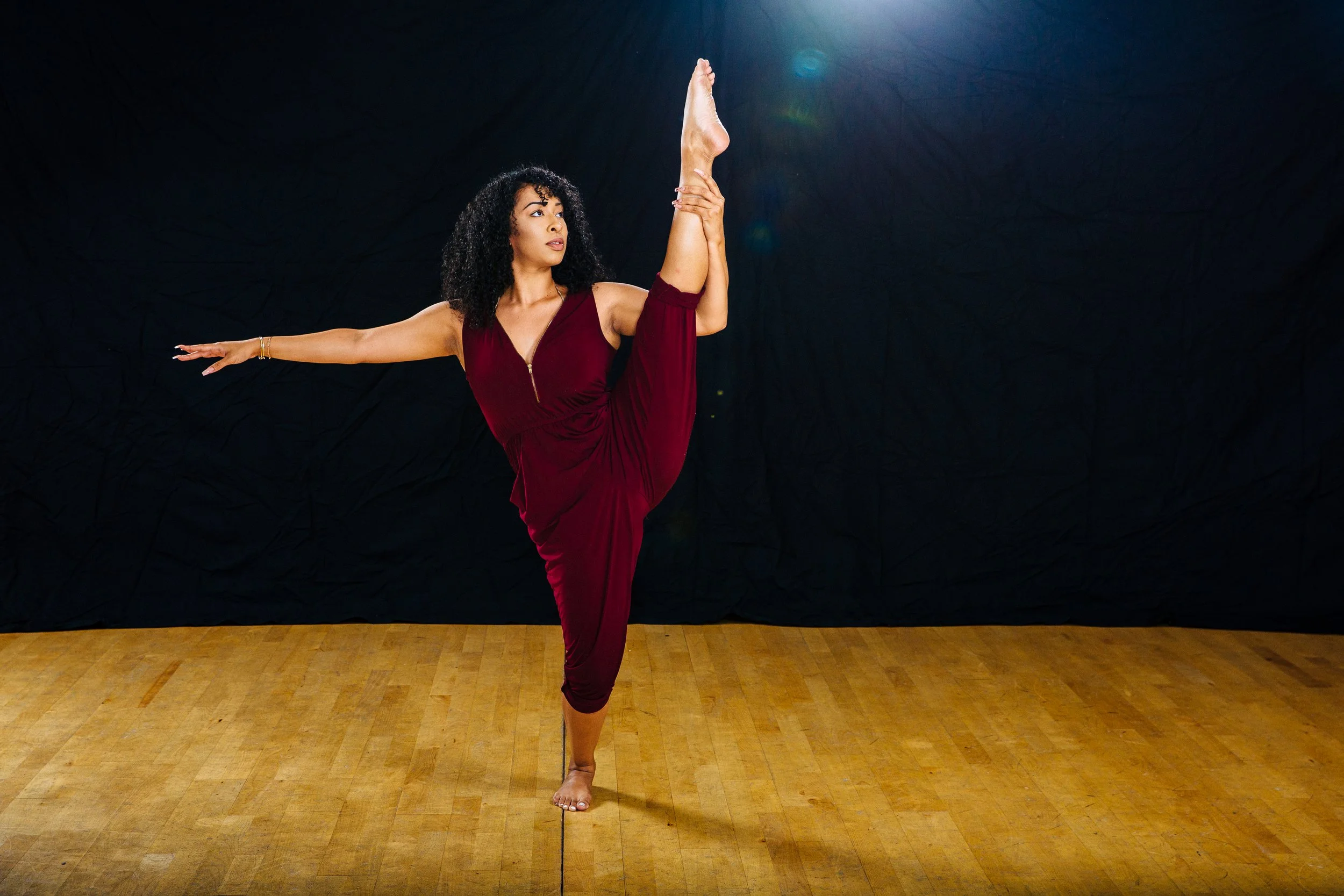 A woman in a red dress performing a yoga pose on a wooden floor against a dark background.