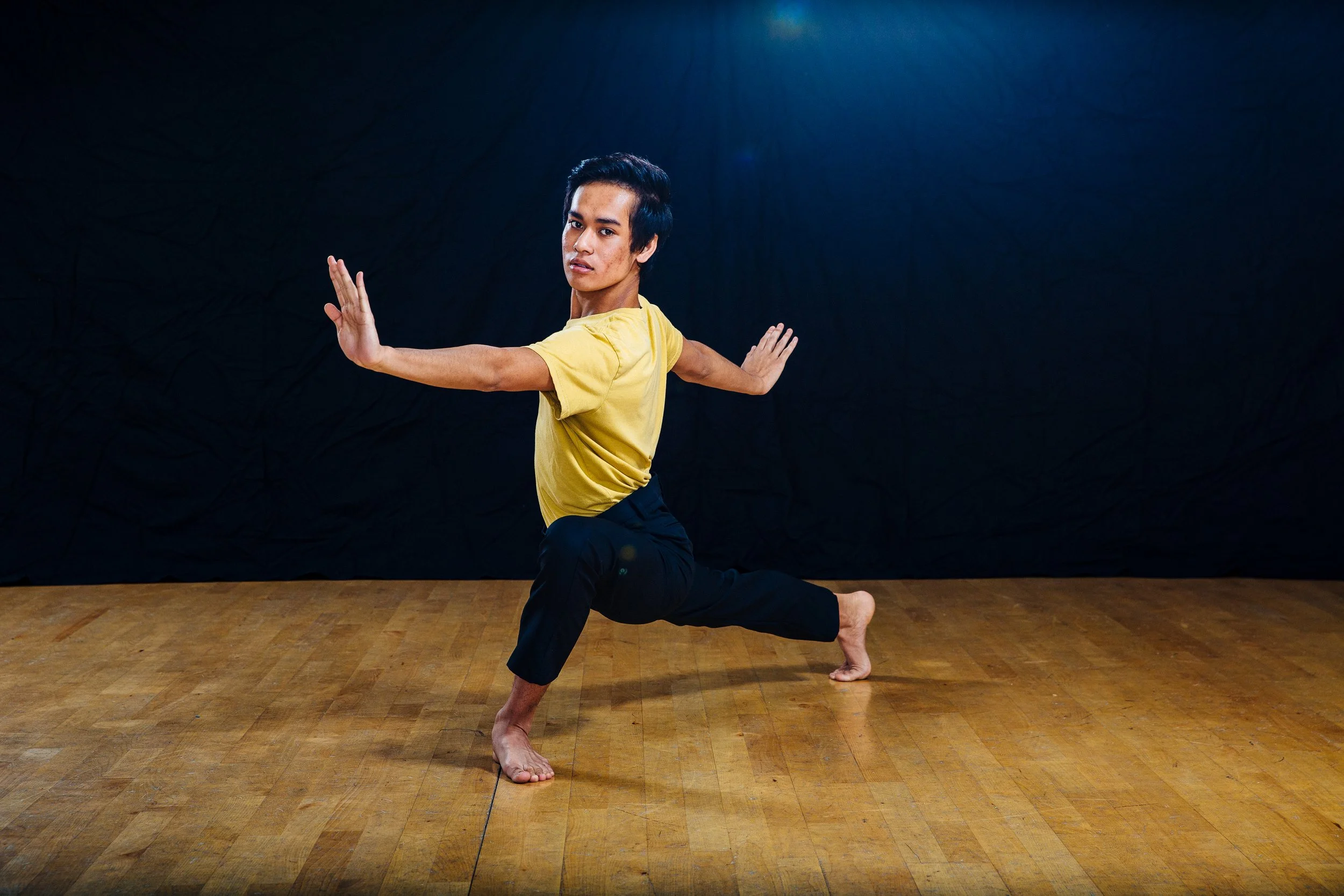 A young man practicing yoga indoors, performing a low lunge pose with his arms extended outward, on a wooden floor against a dark background.