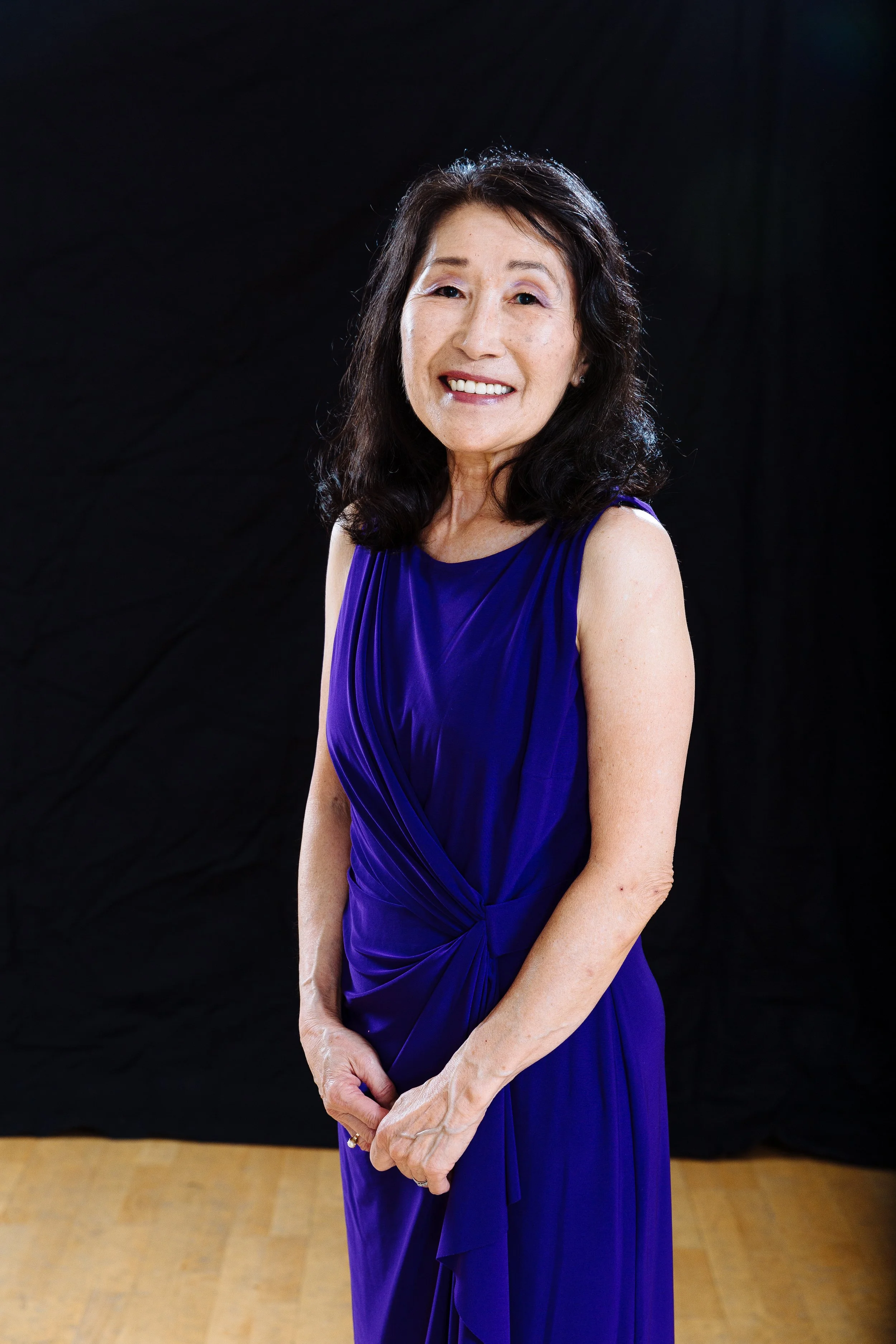 A smiling elderly Asian woman with dark, shoulder-length hair wearing a sleeveless royal blue dress, standing against a black background.