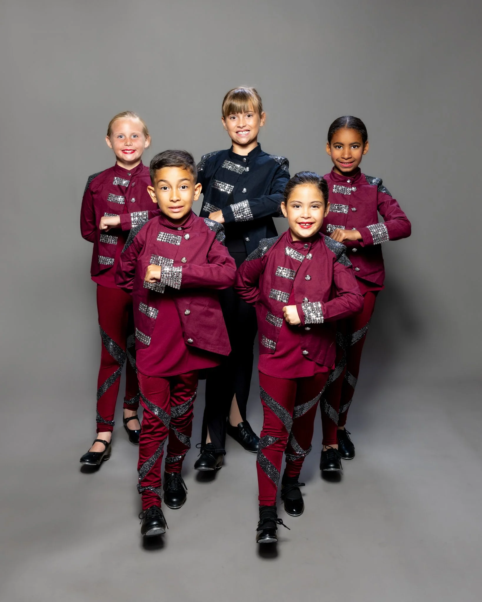 Group of children in matching maroon and black costumes, standing in a formation against a gray background.