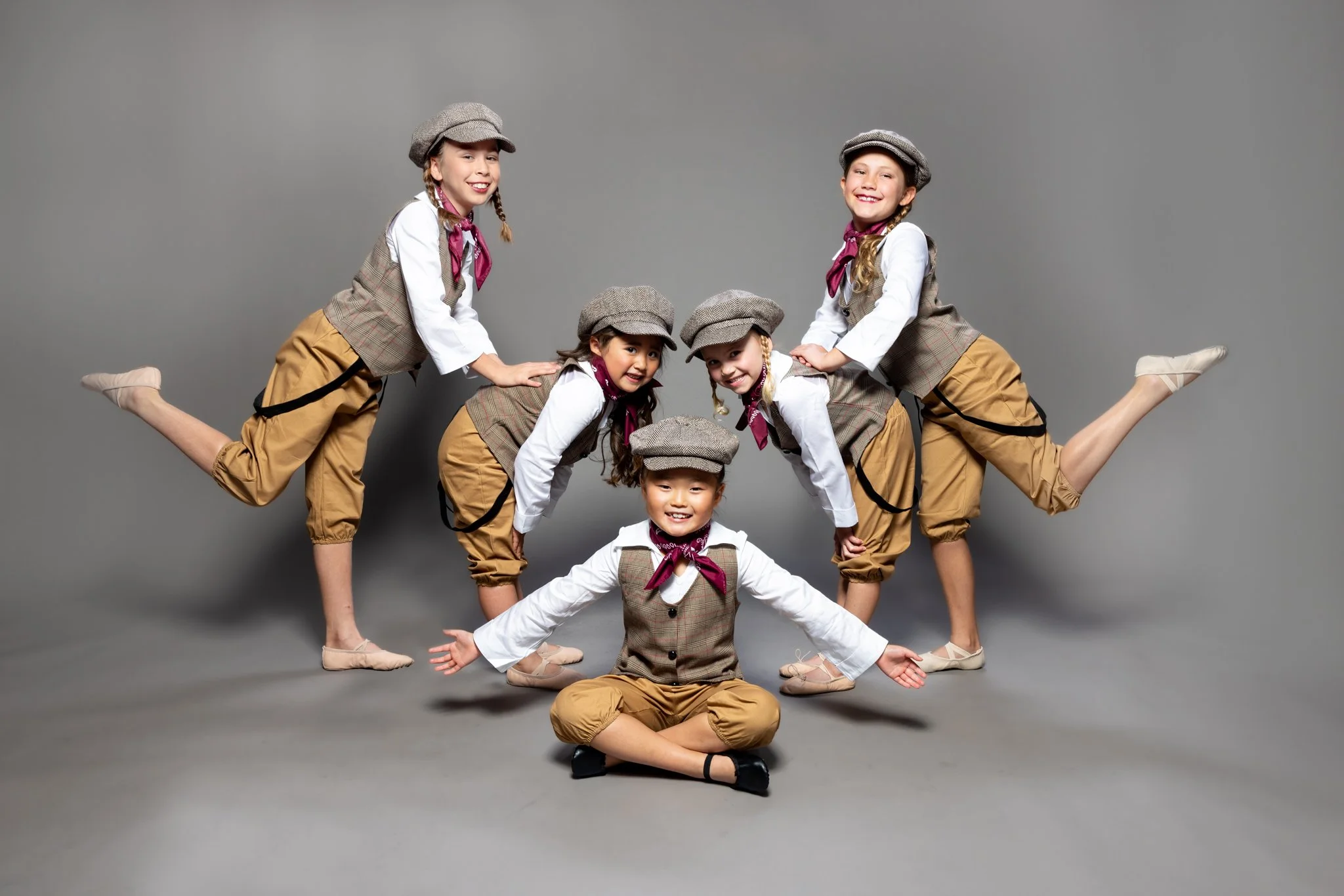 Five young girls dressed in vintage-style costumes, including plaid vests, khaki pants, and matching newsboy caps, performing a coordinated balancing pose against a gray background.