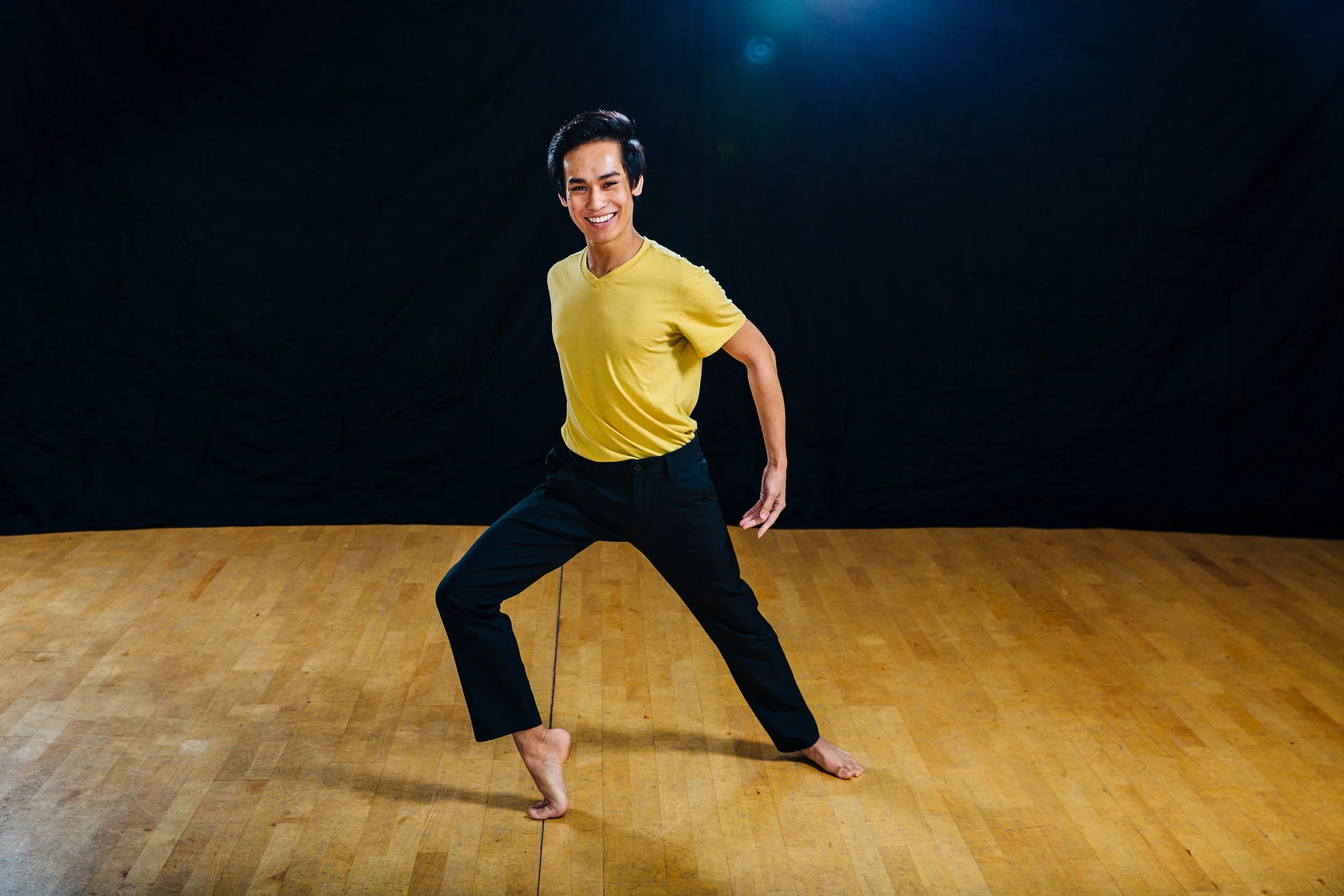 A smiling person with short black hair, wearing a yellow t-shirt and black pants, standing barefoot on a wooden floor against a dark background.