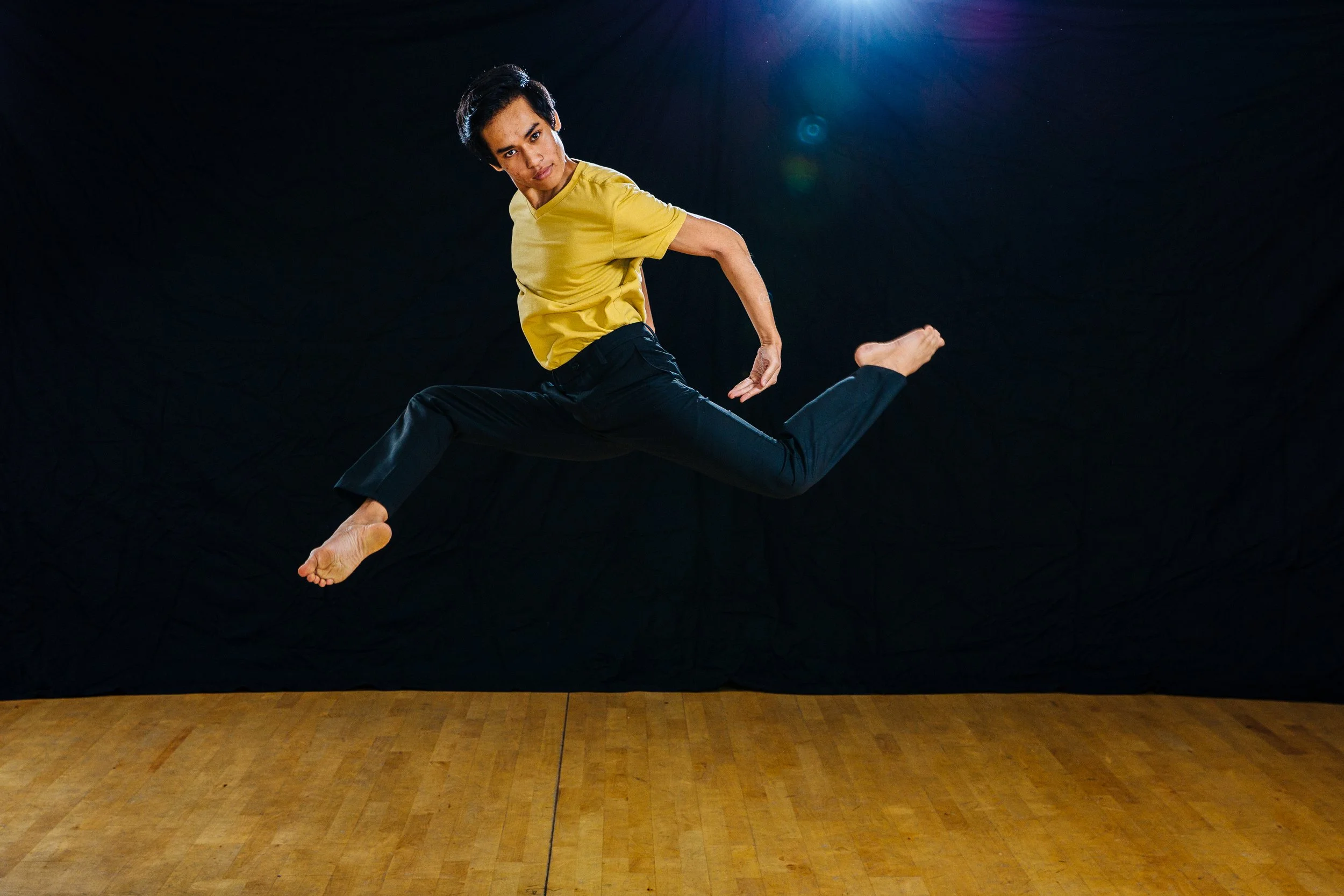 Person mid-air jumping on a wooden floor against a dark backdrop, wearing a yellow shirt and black pants.