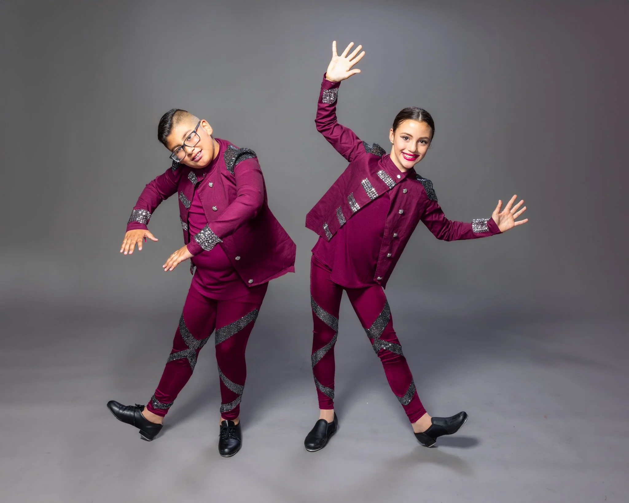 Two young dancers in matching maroon costumes with silver embellishments, dancing against a gray background. The dancer on the right is smiling and has her hands raised, while the dancer on the left is leaning and moving her arms gracefully.