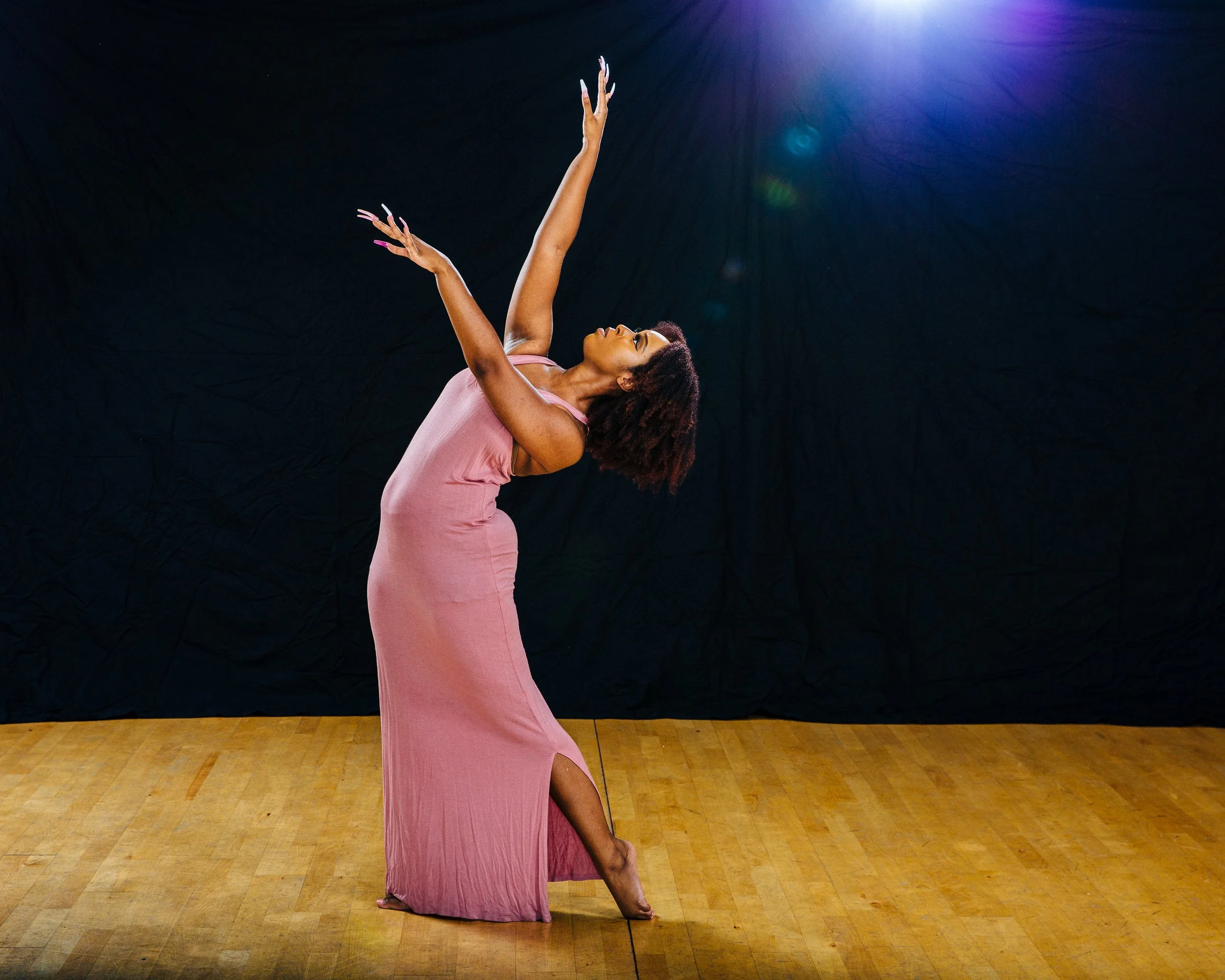 A woman performing a dance pose in a pink dress on a wooden floor with a black background and purple stage light.