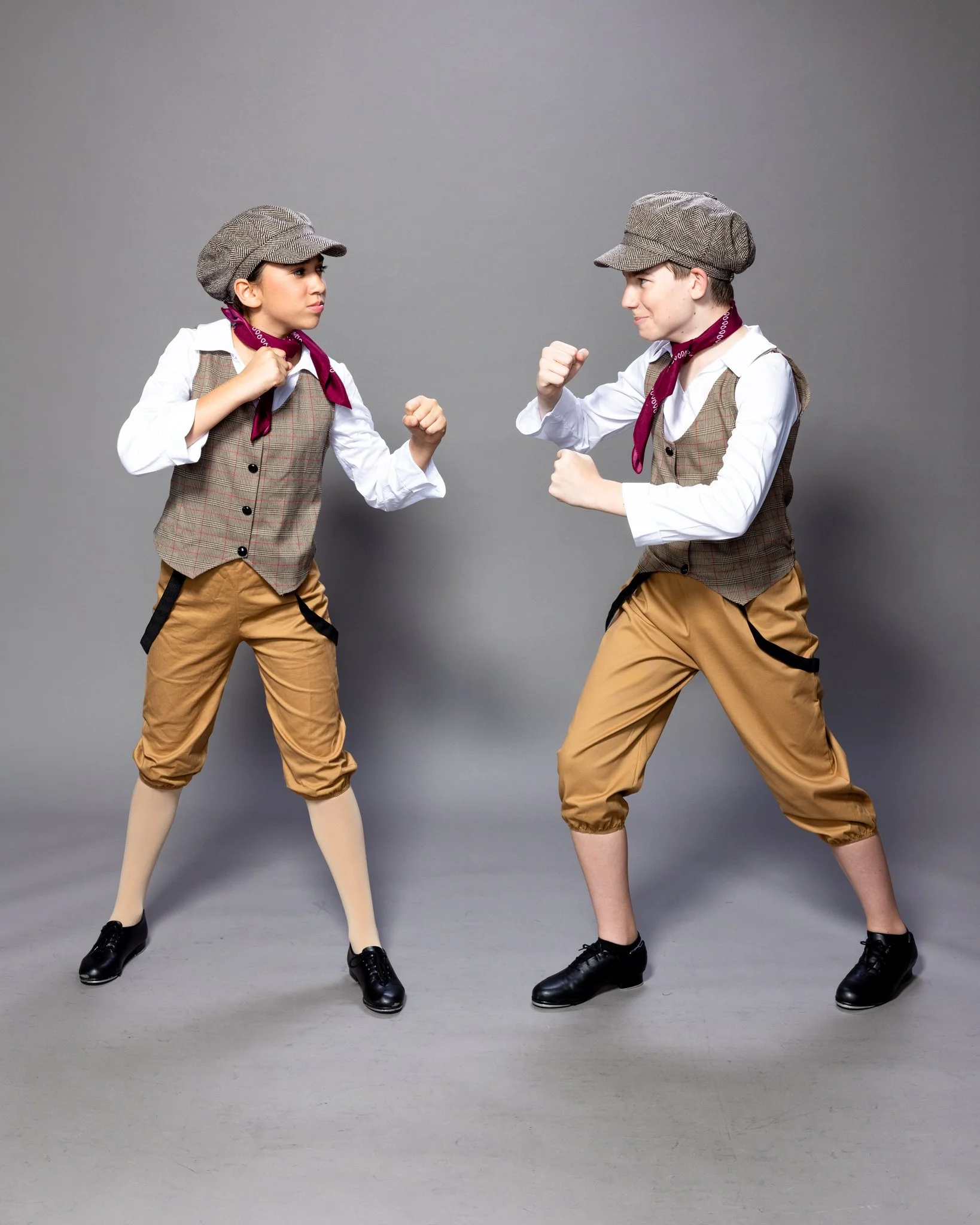Two children dressed as vintage burglars, wearing brown plaid vests, hats, beige pants, white shirts, and maroon scarves, stand facing each other with fists raised as if fighting.