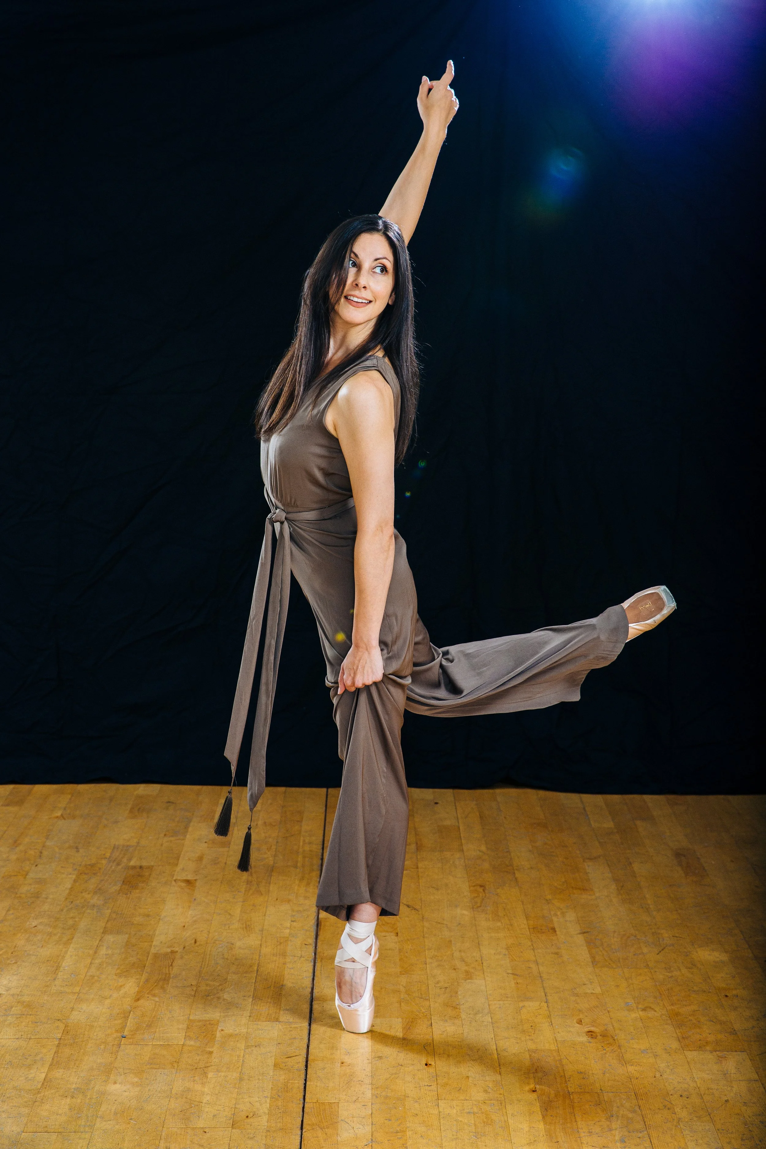 A woman dancing ballet on pointe, wearing a brown jumpsuit and standing on a wooden dance floor against a dark background with a light source overhead.