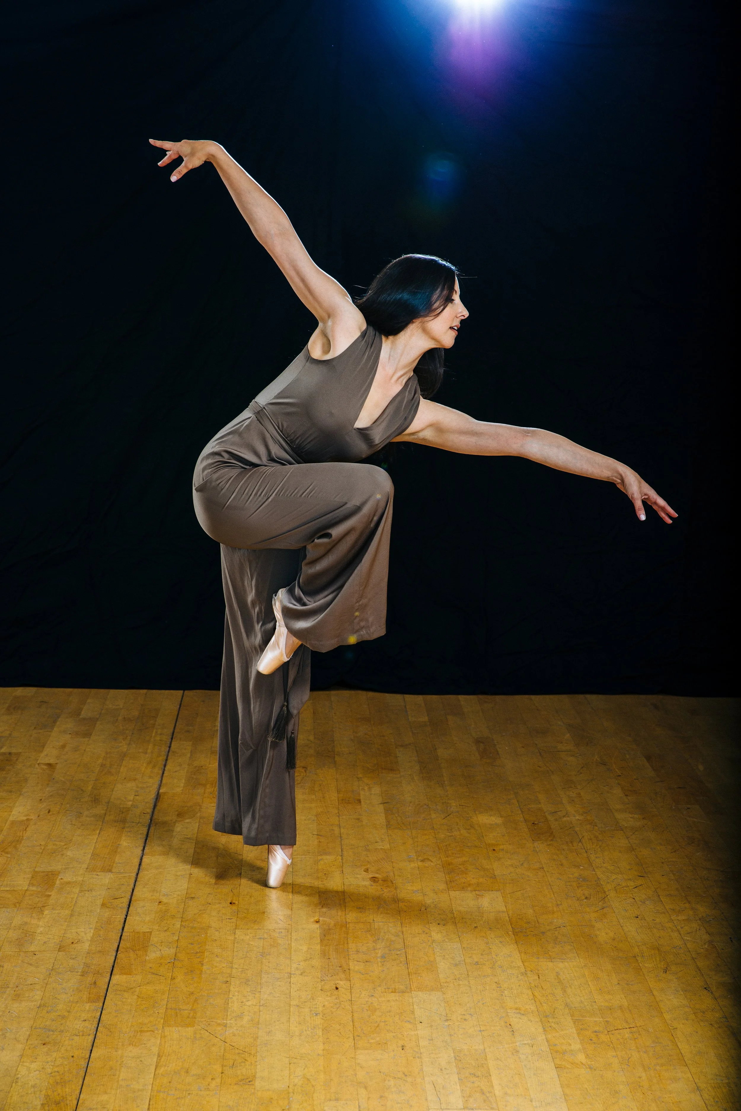 A woman in a taupe jumpsuit performing a ballet move on pointe, with one arm raised and the other extended to the side, against a black background with a purple spotlight shining from above.