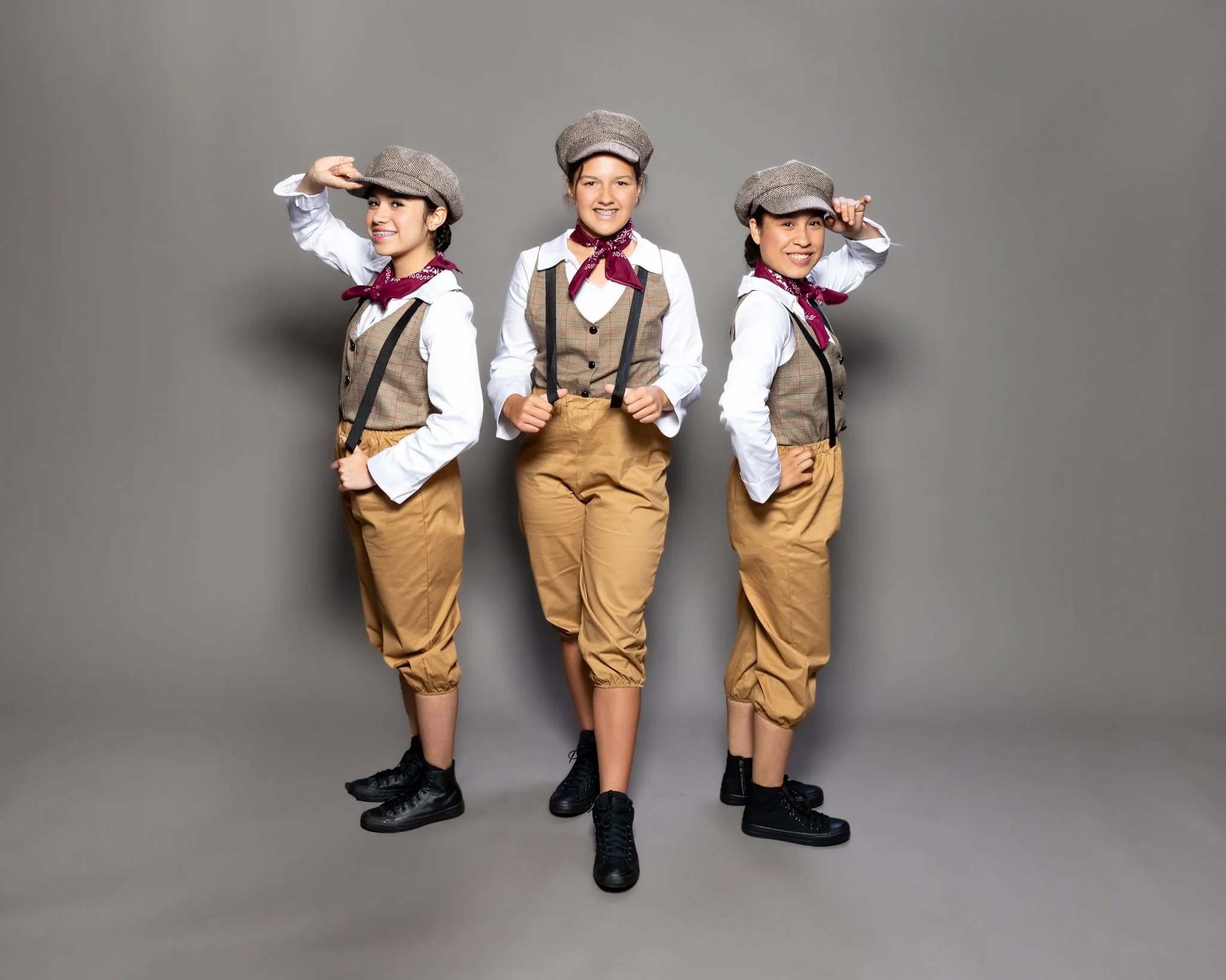 Three young girls dressed as vintage detectives or reporters, wearing beige plaid vests, khaki pants, white shirts, maroon neckerchiefs, gray flat caps, and black sneakers, standing against a gray background with confident poses.