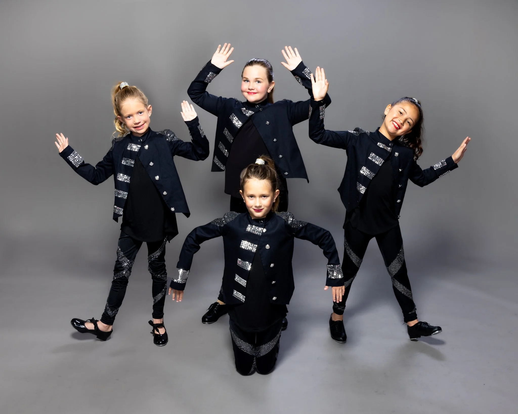 Five young girls in matching black dance costumes with sparkly silver accents, posing in a dance studio with a gray background.