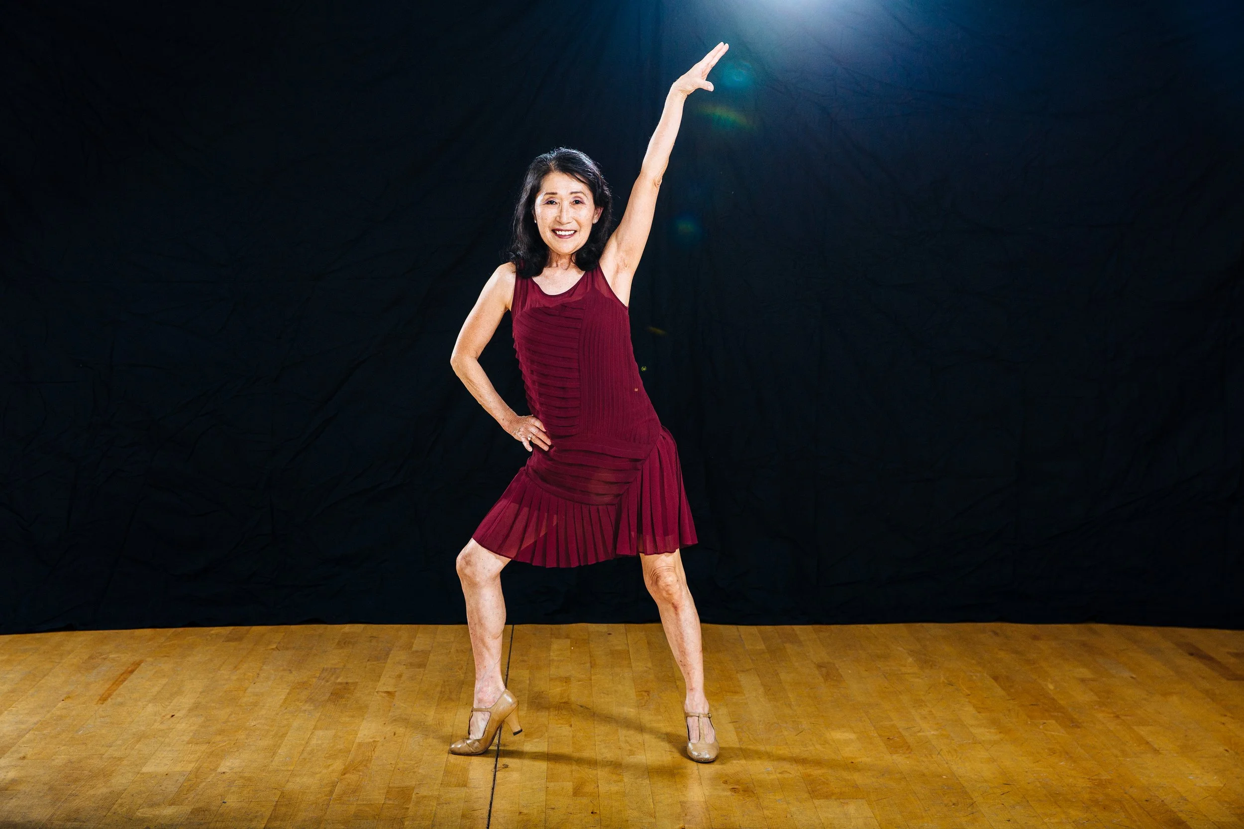 A woman standing on a wooden floor with a black background, wearing a red dress and high heels, striking a pose with one arm raised and smiling.