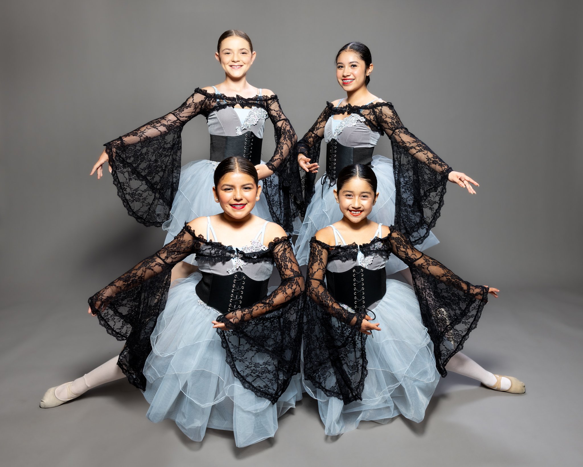 Four young girls dressed in ballet costumes with black lace and gray skirts, smiling and posing in a dance studio.