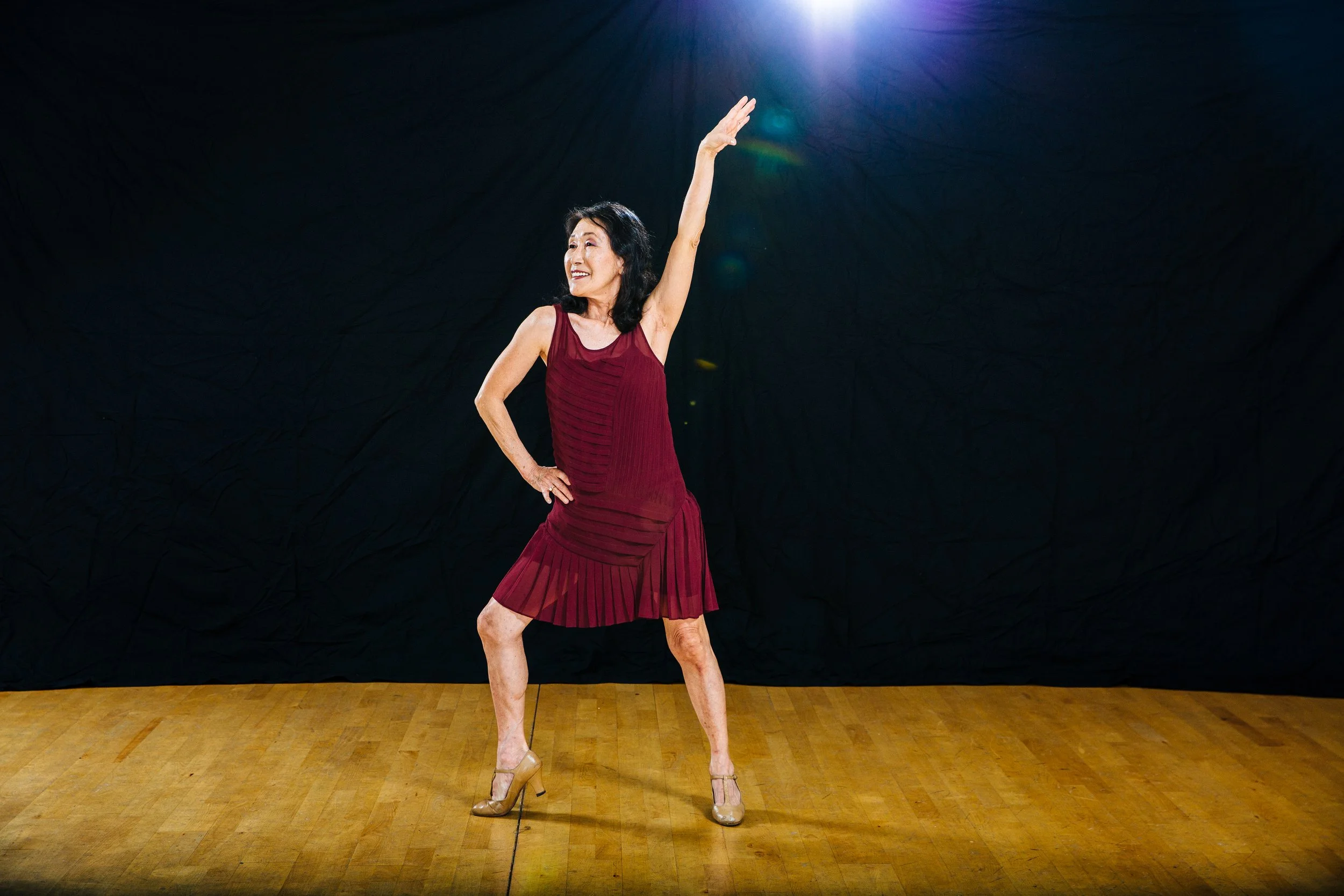 Woman in a red dress dancing on a wooden floor with a black background and light shining down from above.