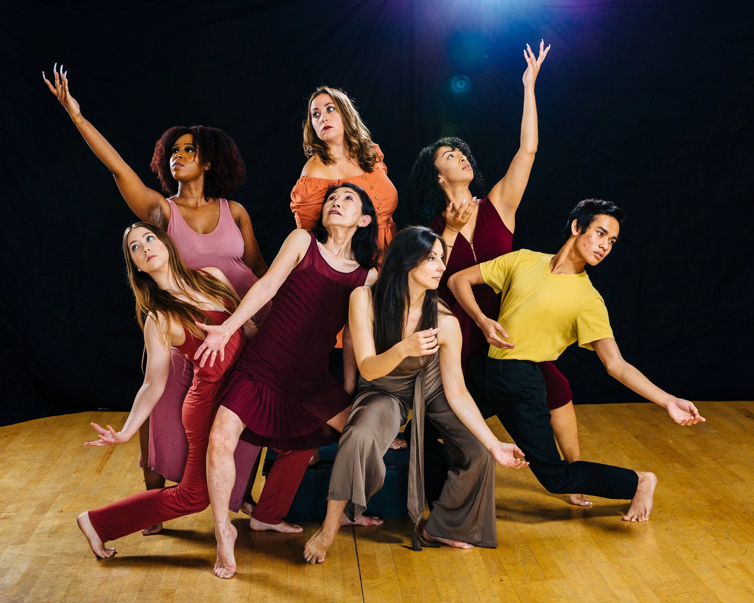 A diverse group of seven women performing a theatrical or dance pose on stage with a black backdrop and wooden floor.