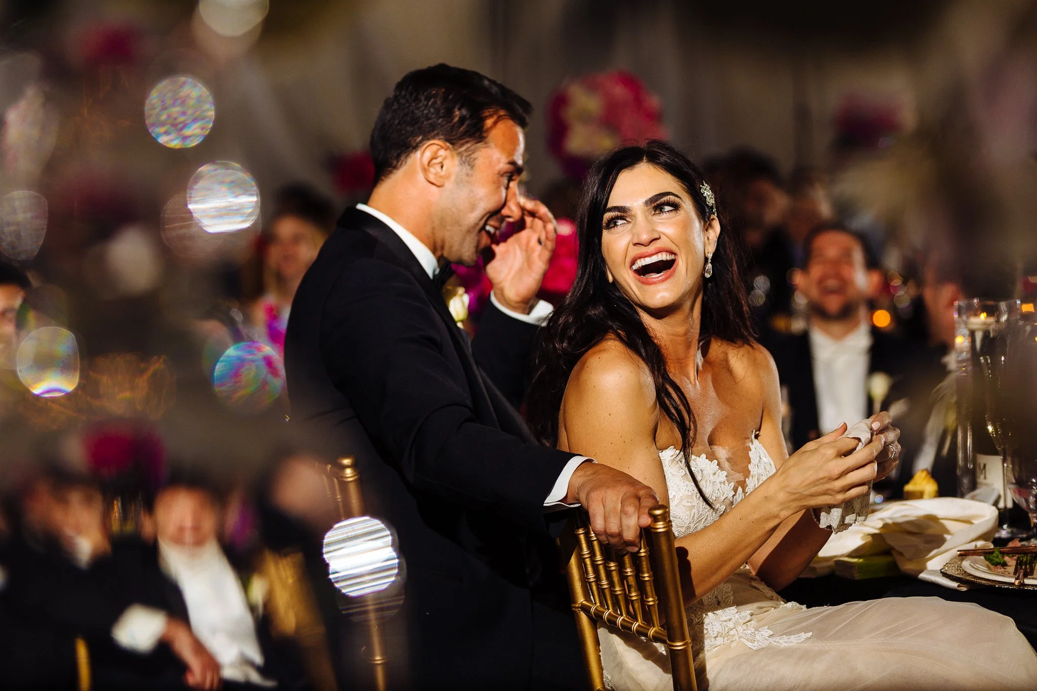 A bride and groom sitting at a wedding reception, the bride laughing and the groom smiling. The woman has long dark hair, a white lace dress, and elegant earrings. The man is in a black suit with a white shirt. The background is filled with blurred g