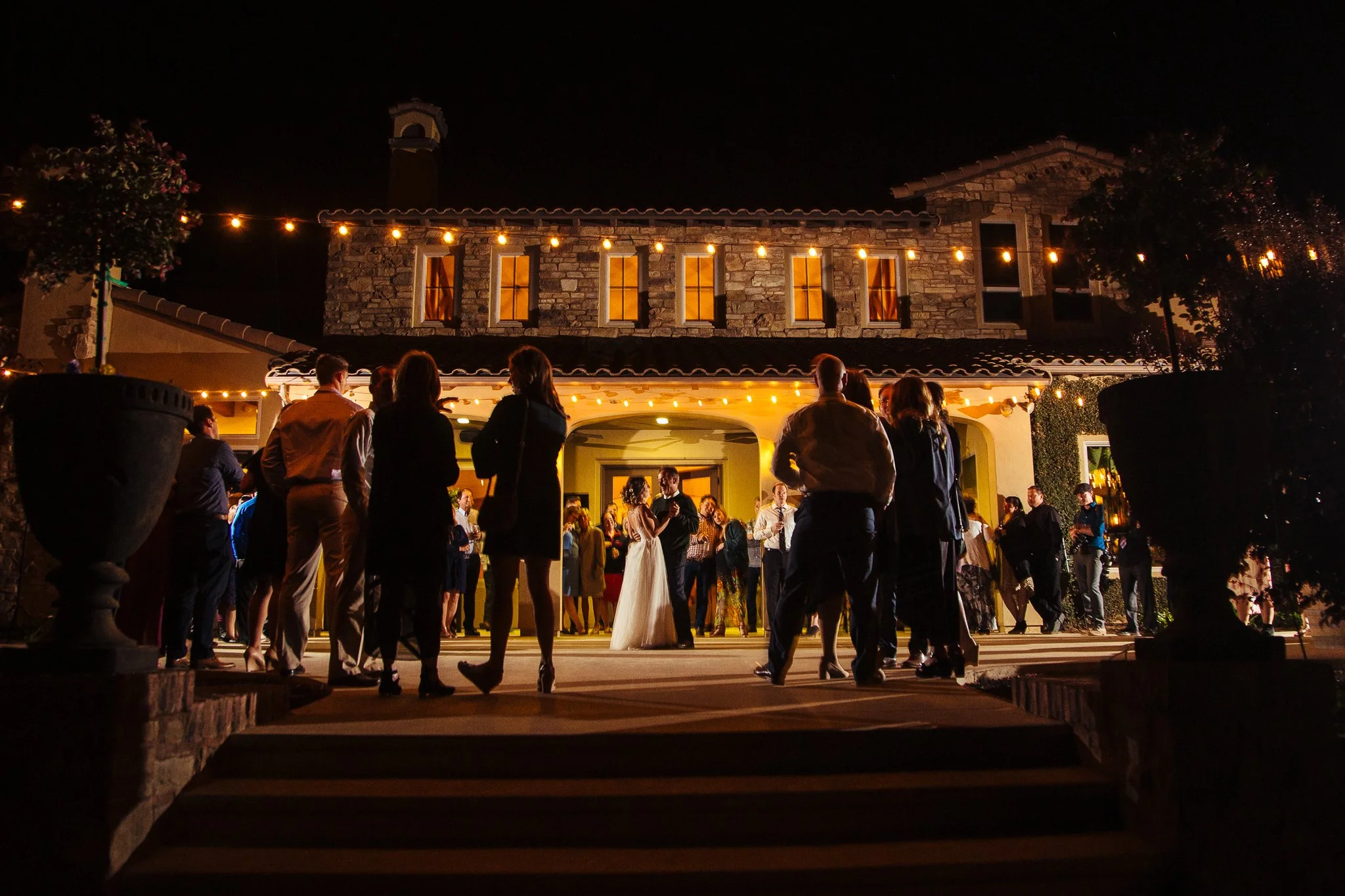 People gathered outside a lit house at night, possibly for a celebration or wedding, with string lights hanging overhead and a stone and stucco exterior.