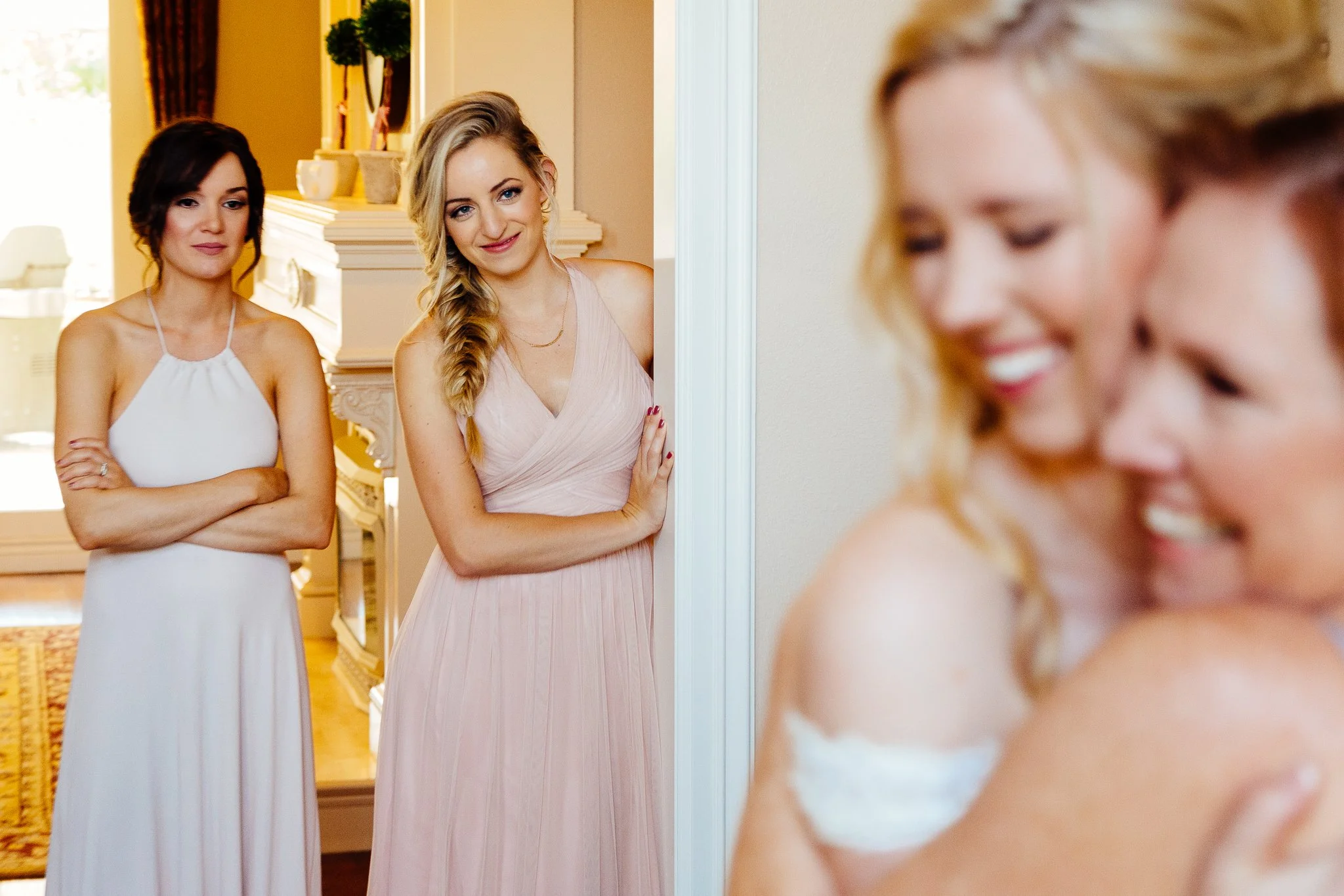 Three women, one with dark hair and wearing a white dress, and another with blonde hair in a pink dress, are smiling and looking at a bride who is hugging a woman with reddish hair. The scene appears to be indoors, in a warmly lit room.