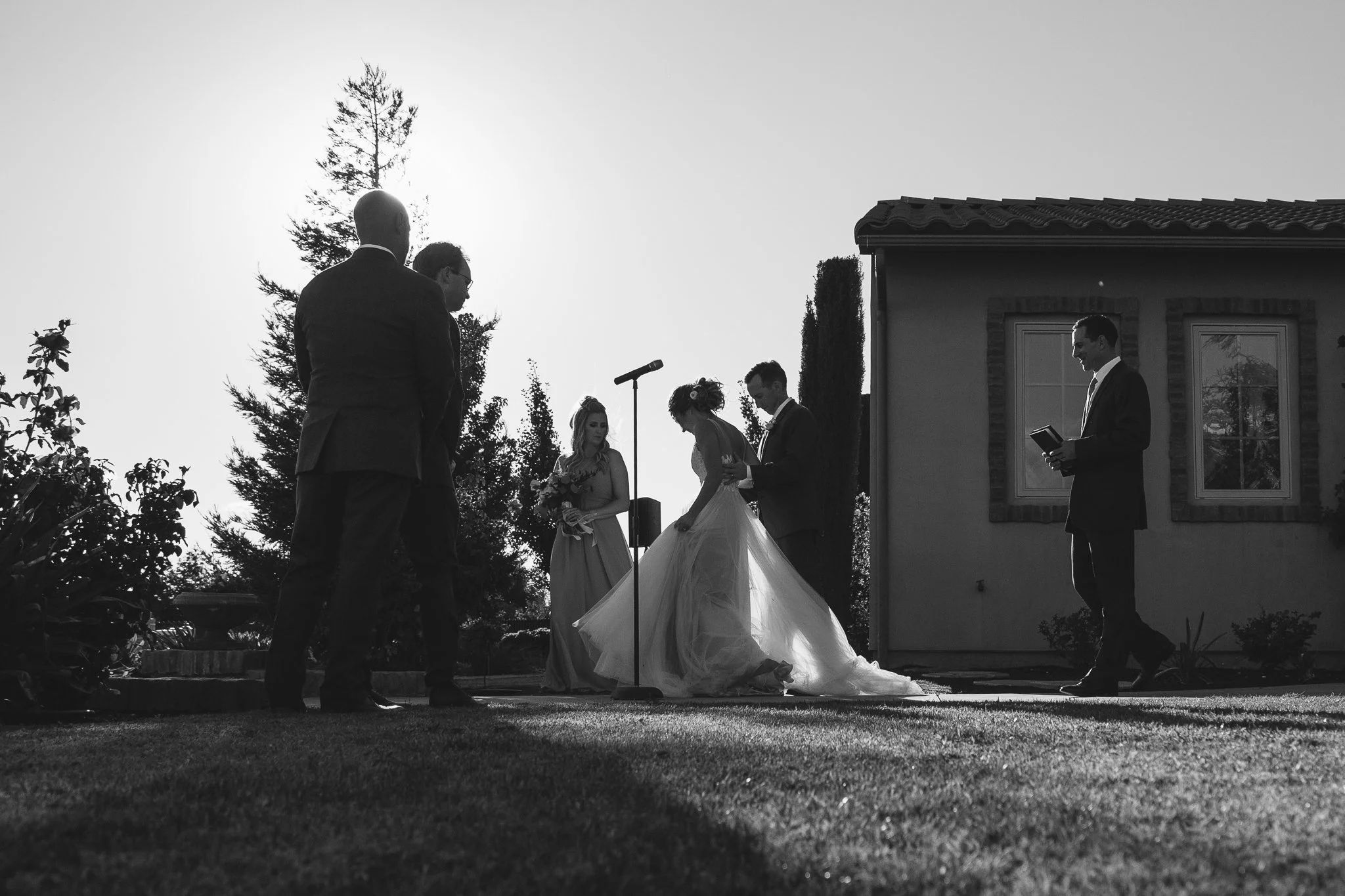 A black and white photo of a wedding ceremony outdoors with the bride, groom, and officiant, along with guests, with a house in the background and trees.