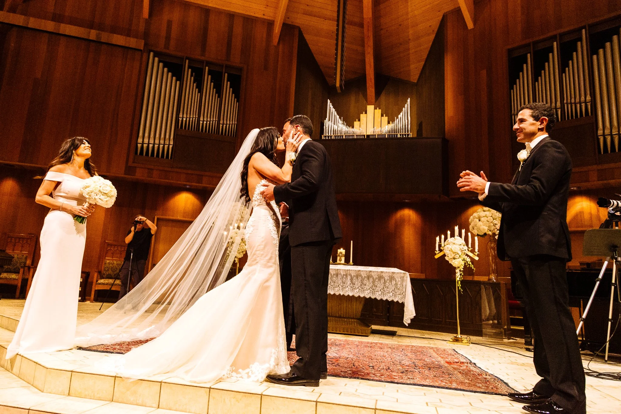 A couple kisses during their wedding ceremony in a church with wood-paneled walls and organ pipes, with a woman holding a bouquet and a man clapping nearby.