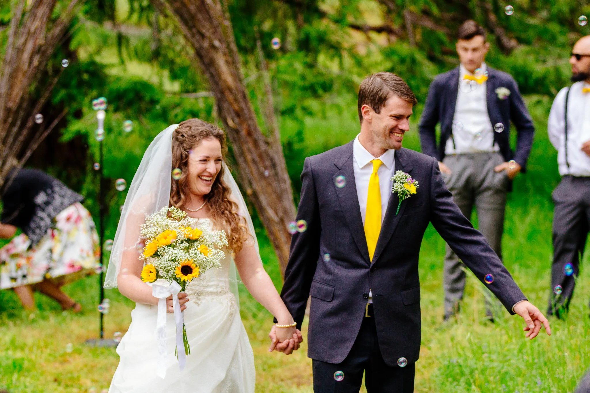 A bride and groom holding hands and smiling during their outdoor wedding ceremony, surrounded by groomsmen and greenery.