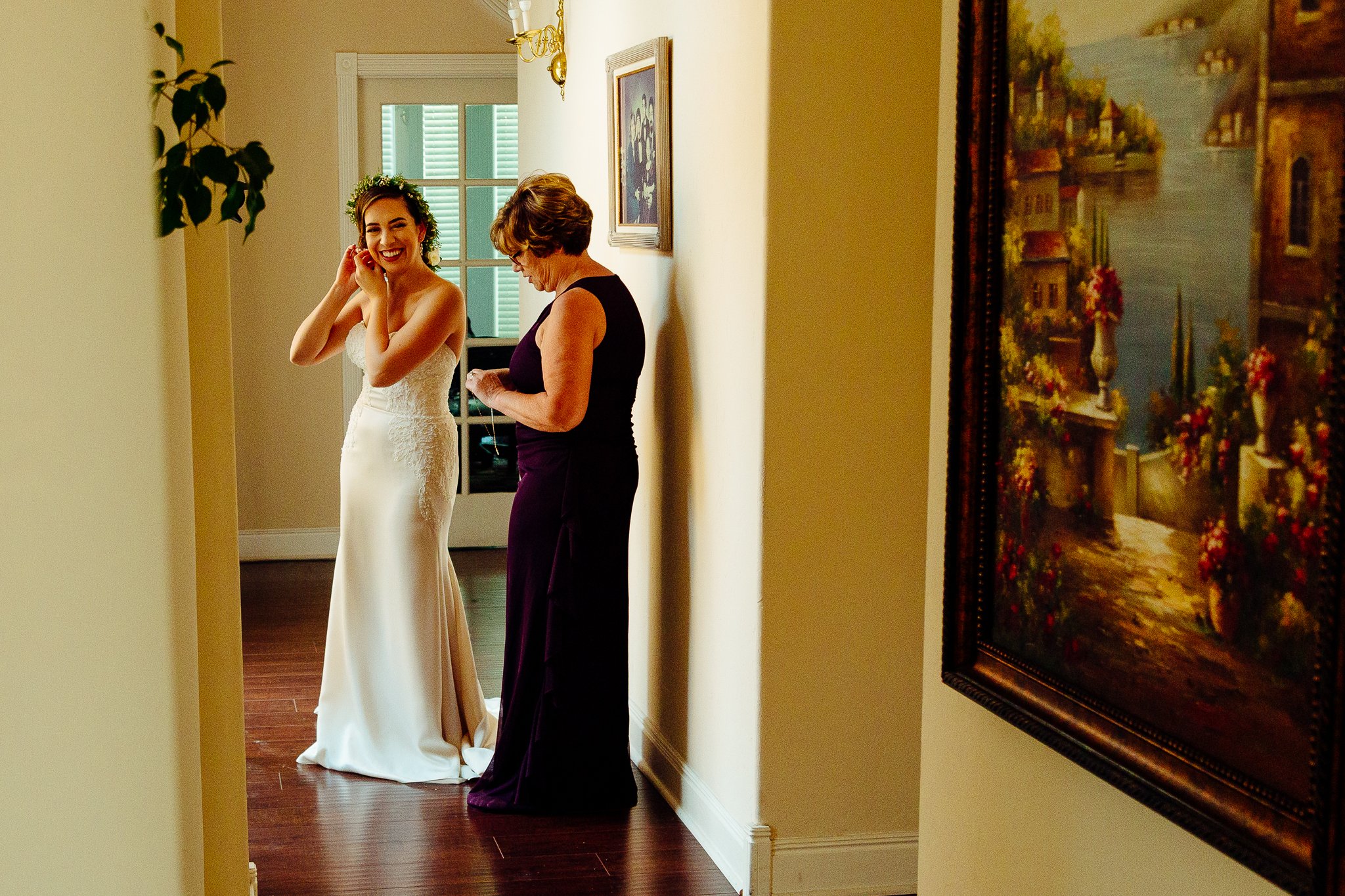 A bride is smiling while talking on her phone, standing next to an older woman in a purple dress in a hallway decorated with paintings and a window.