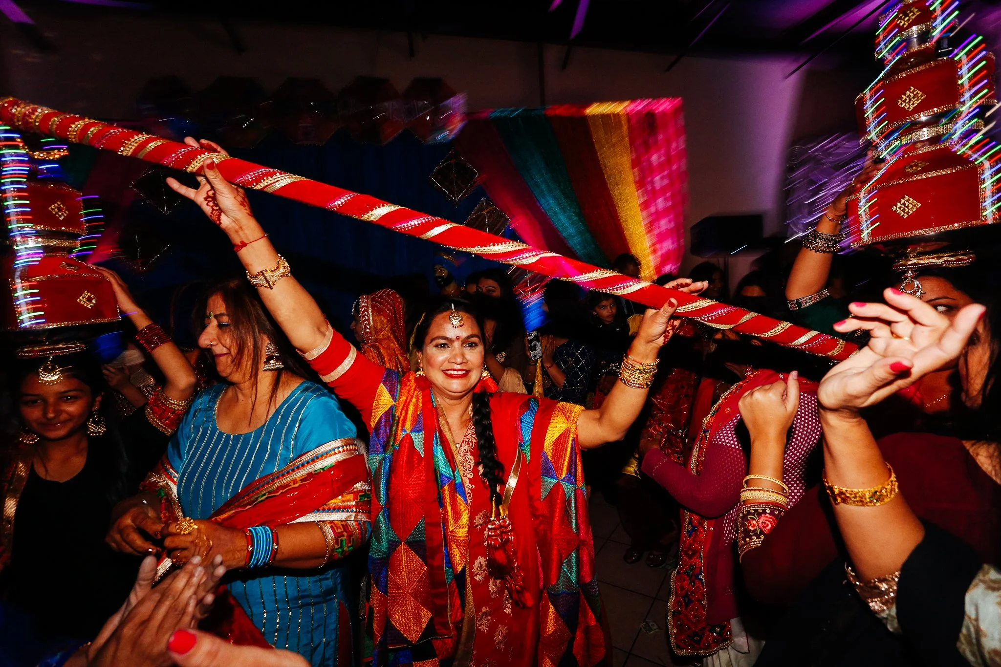 Women celebrating at a colorful Indian festival, dressed in traditional attire, holding decorated pots and a long stick, with vibrant decorations and lights in the background.