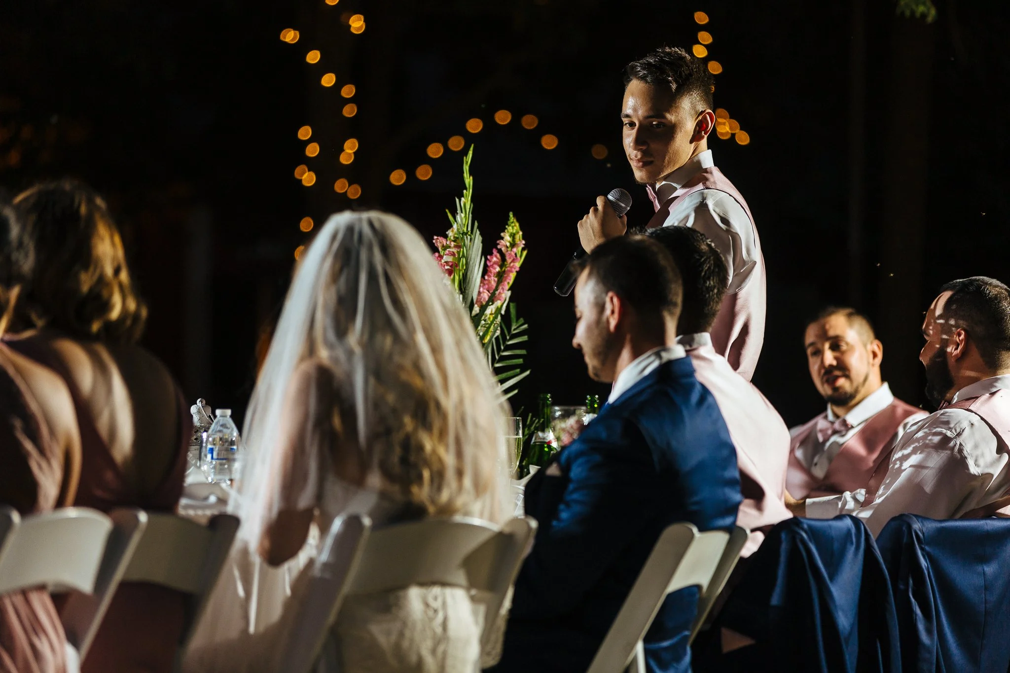 A man giving a speech with a microphone at a wedding reception, with a bride in a white dress and veil and other guests seated around the table, decorated with flowers and string lights in the background.