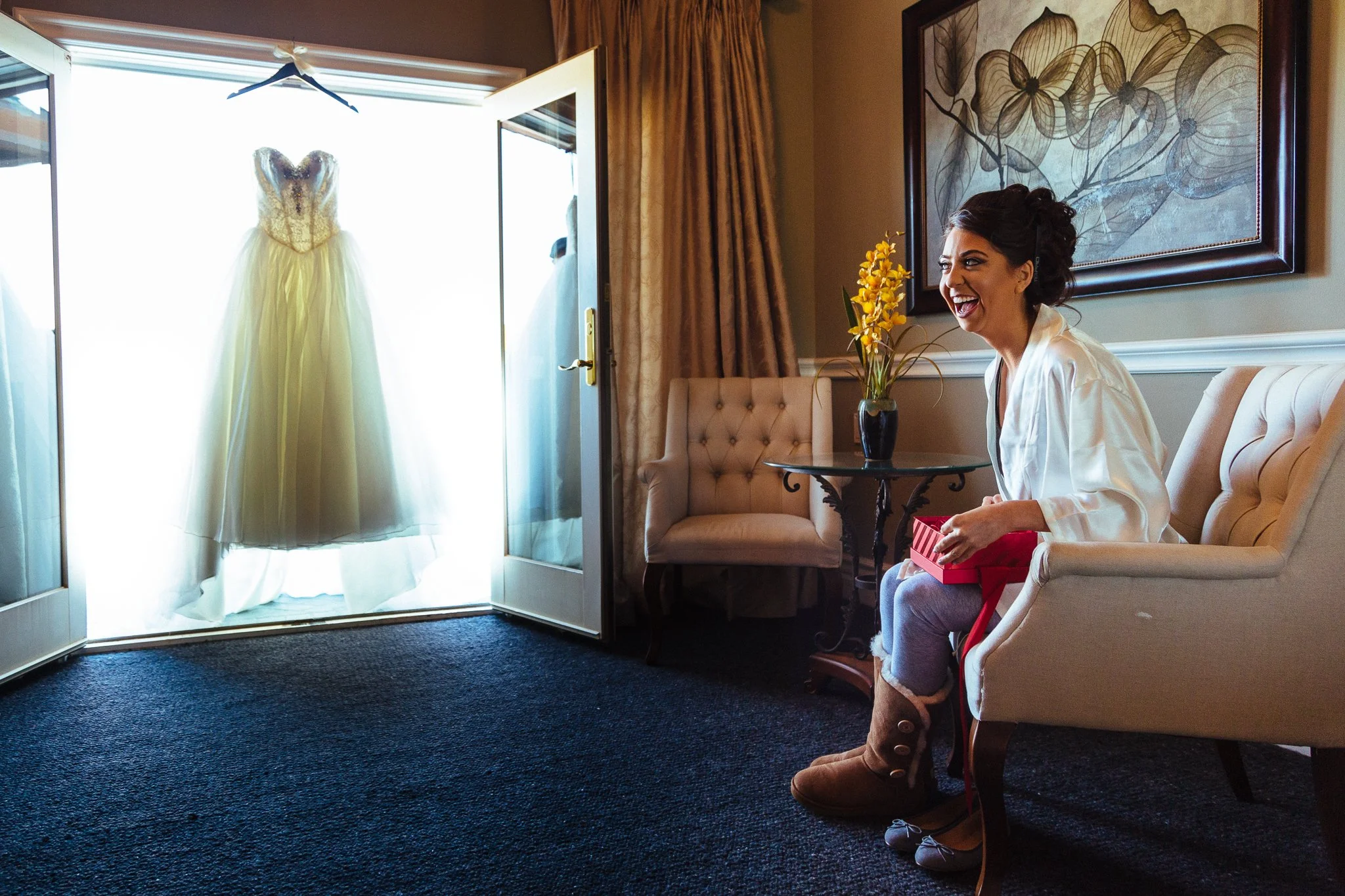 A woman sitting on a beige upholstered chair, smiling and holding a small red gift box, in a room with a vase of yellow flowers on a small table, a large painting on the wall, and a wedding dress hanging in the open door to the balcony.
