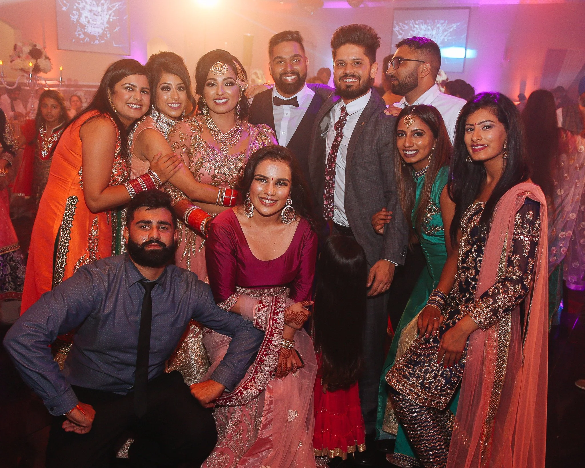 Group of people dressed in traditional Indian attire at a celebration or wedding party, smiling and posing for a photo in a decorated indoor venue.