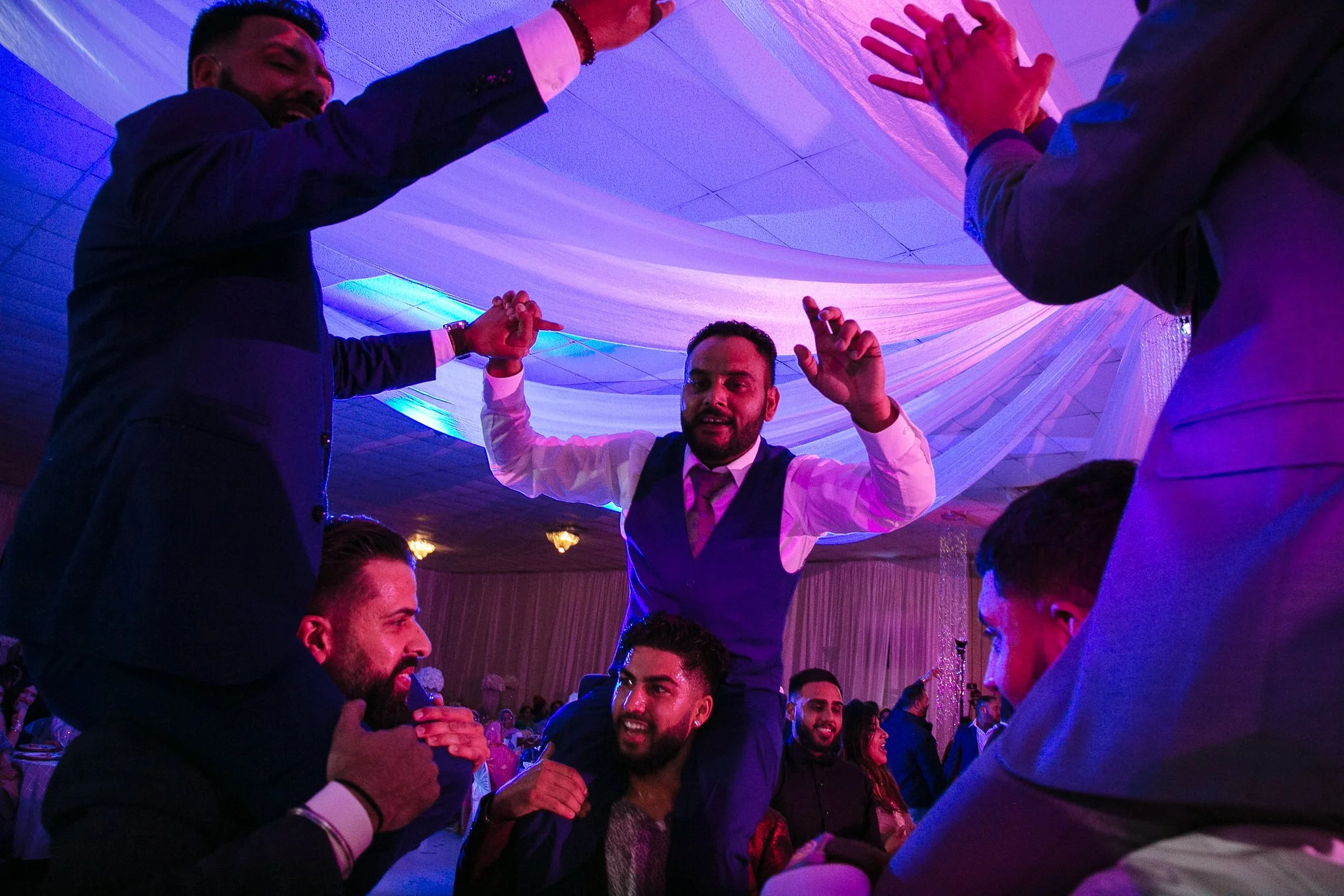 Group of men celebrating at a party, with one man sitting on another's shoulders, raising his hands, amid colorful lighting and draped ceiling decorations.
