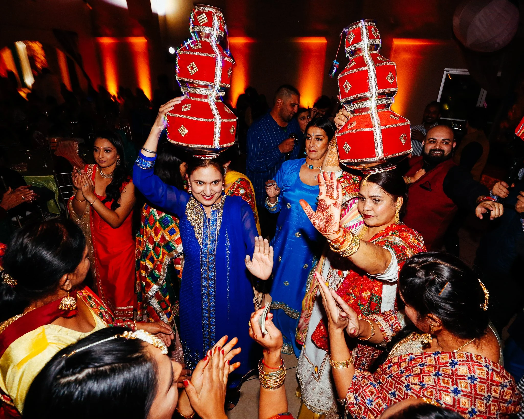 Indian women dressed in colorful traditional attire, performing a dance at a celebration or wedding, with some balancing decorated red pots on their heads, in a lively indoor setting with warm lighting.