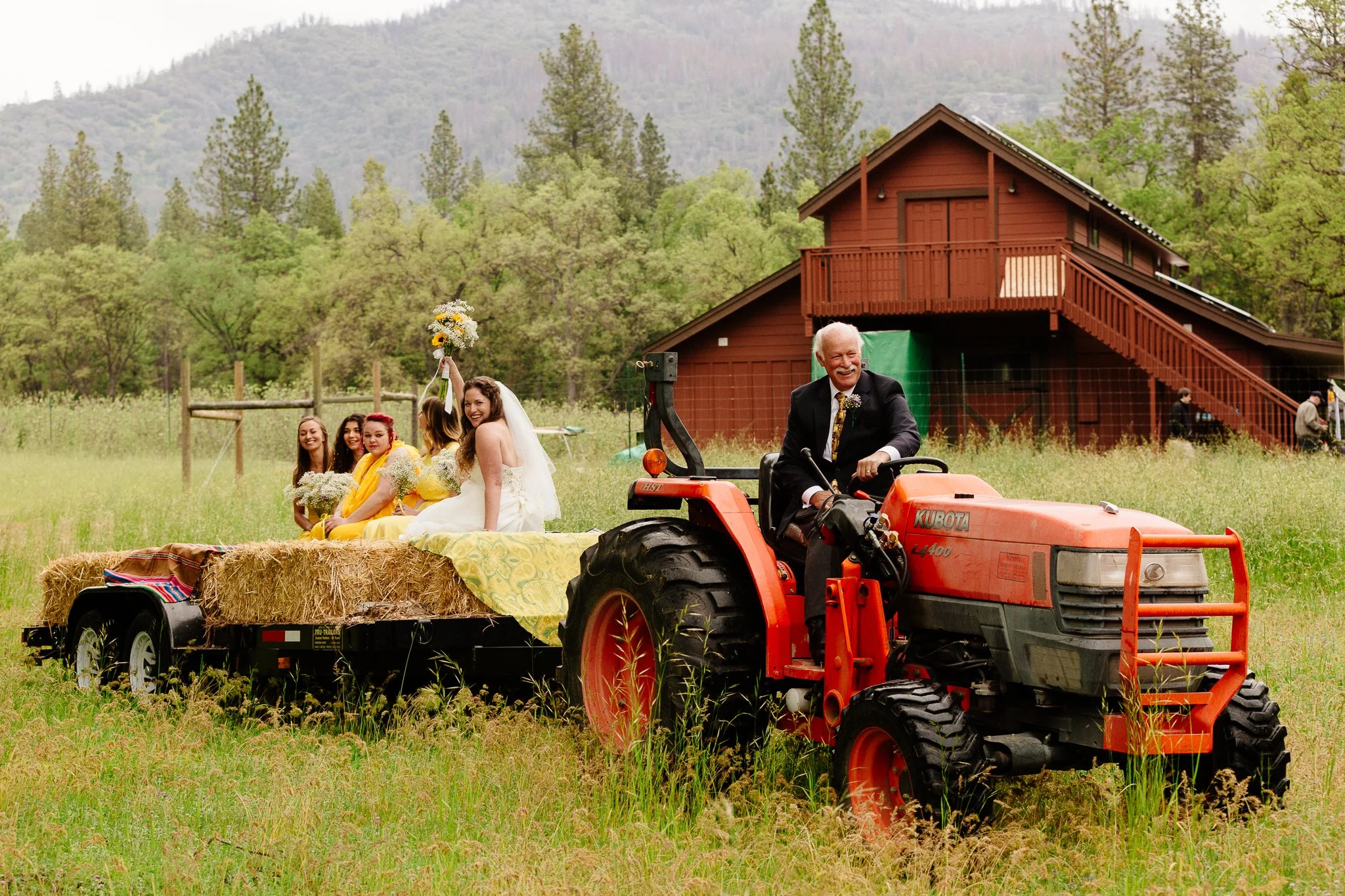 A wedding celebration with a bride and bridesmaids on a hay-filled trailer being pulled by a red tractor in a field, with a brown barn and green trees in the background.